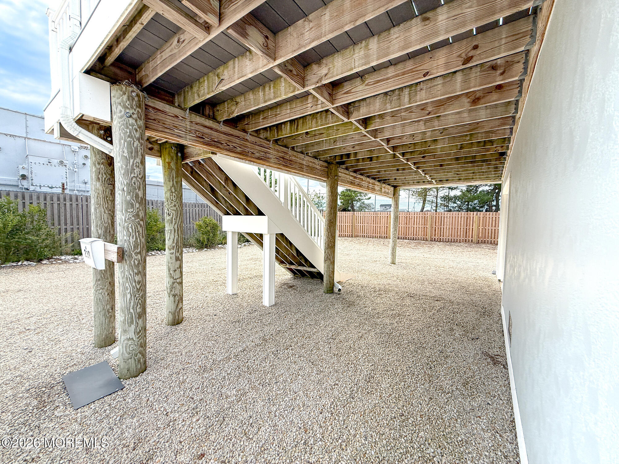 Gravel yard beneath an elevated home, with exposed deck supports, stairs, and a wooden privacy fence.