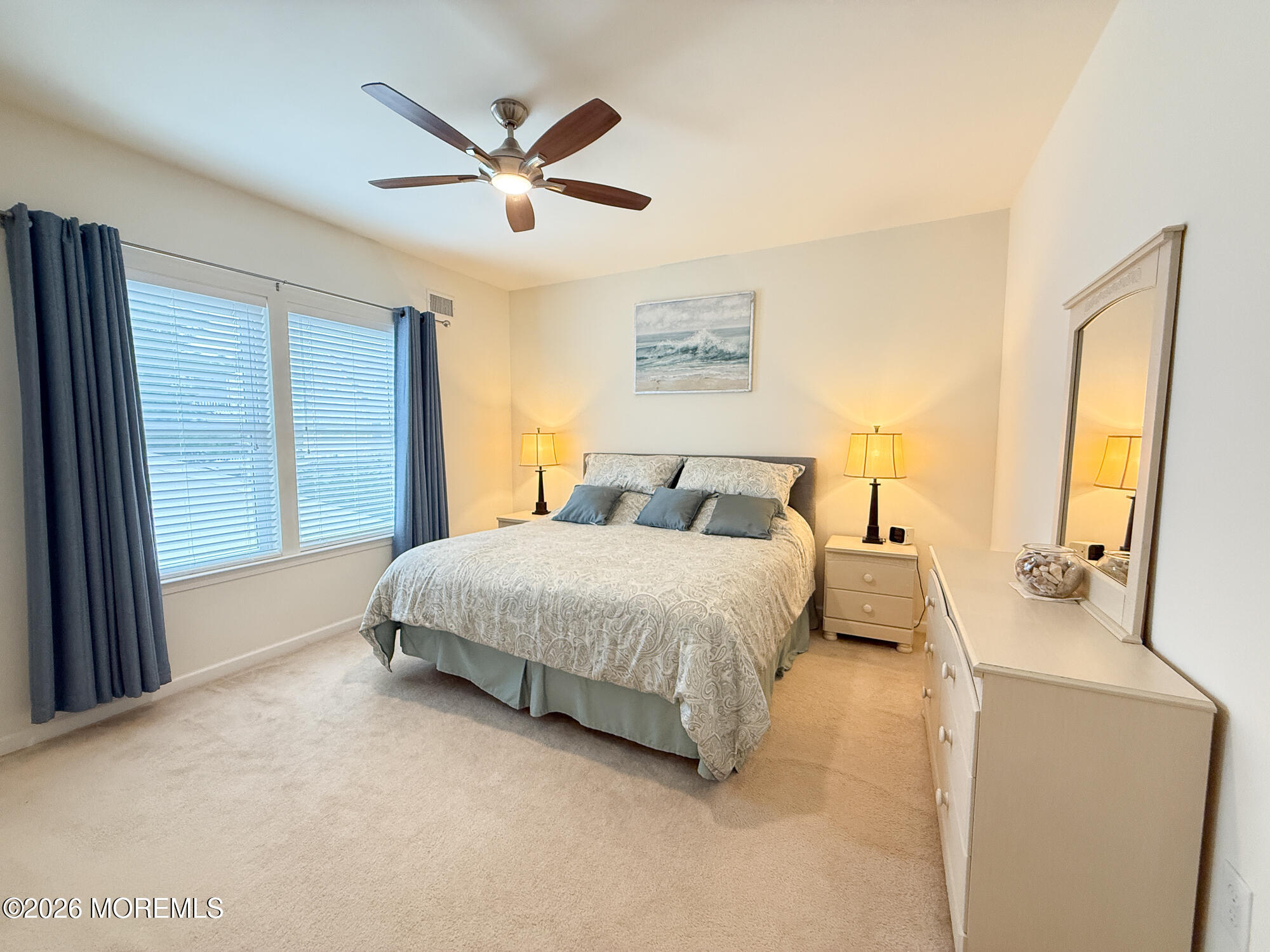 Furnished bedroom with patterned bedding, blue-gray curtains, bedside lamps, and a dresser with mirror.