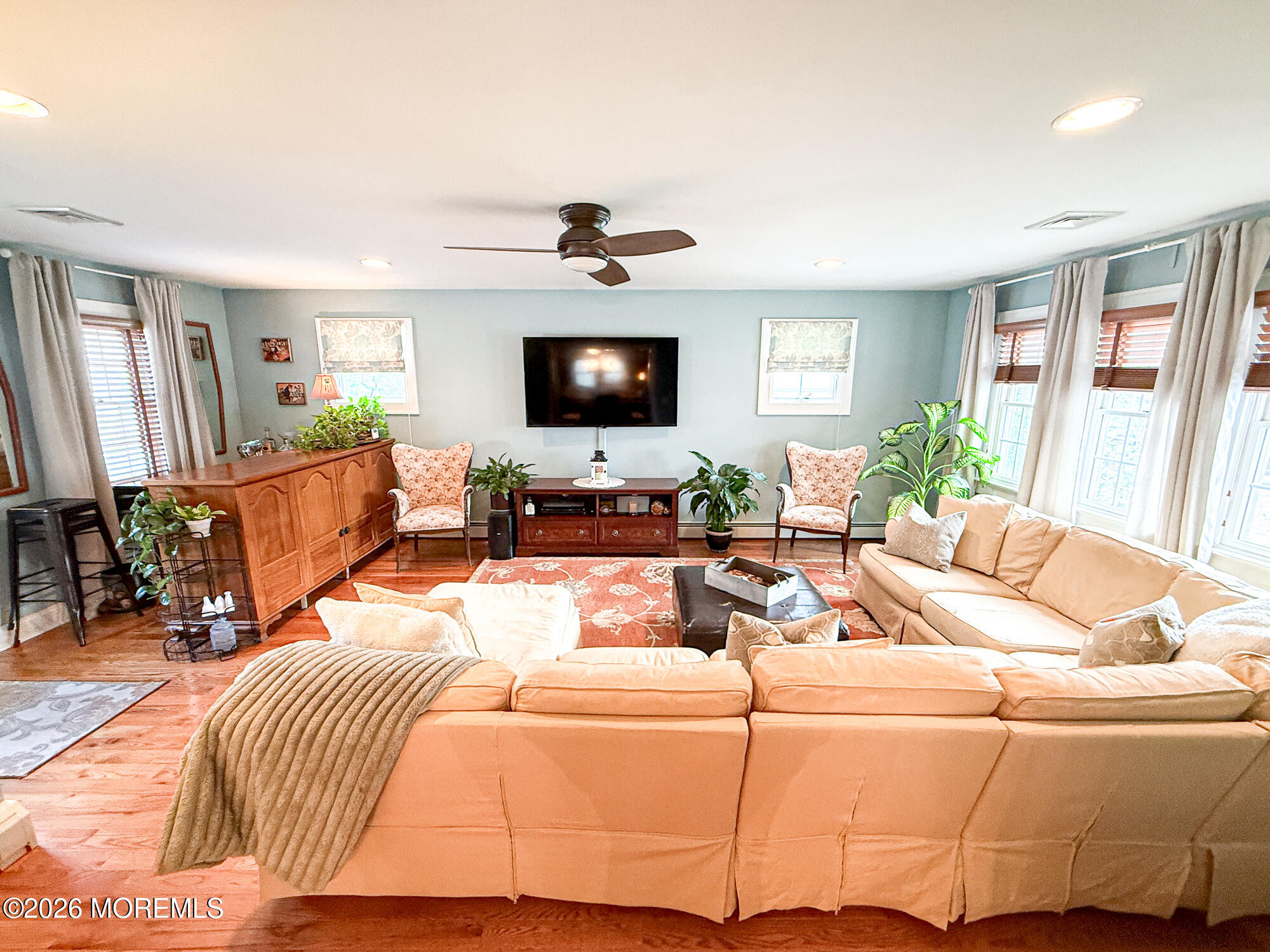 Living room with a peach sectional, wall-mounted television, floral armchairs, and broad curtained windows.