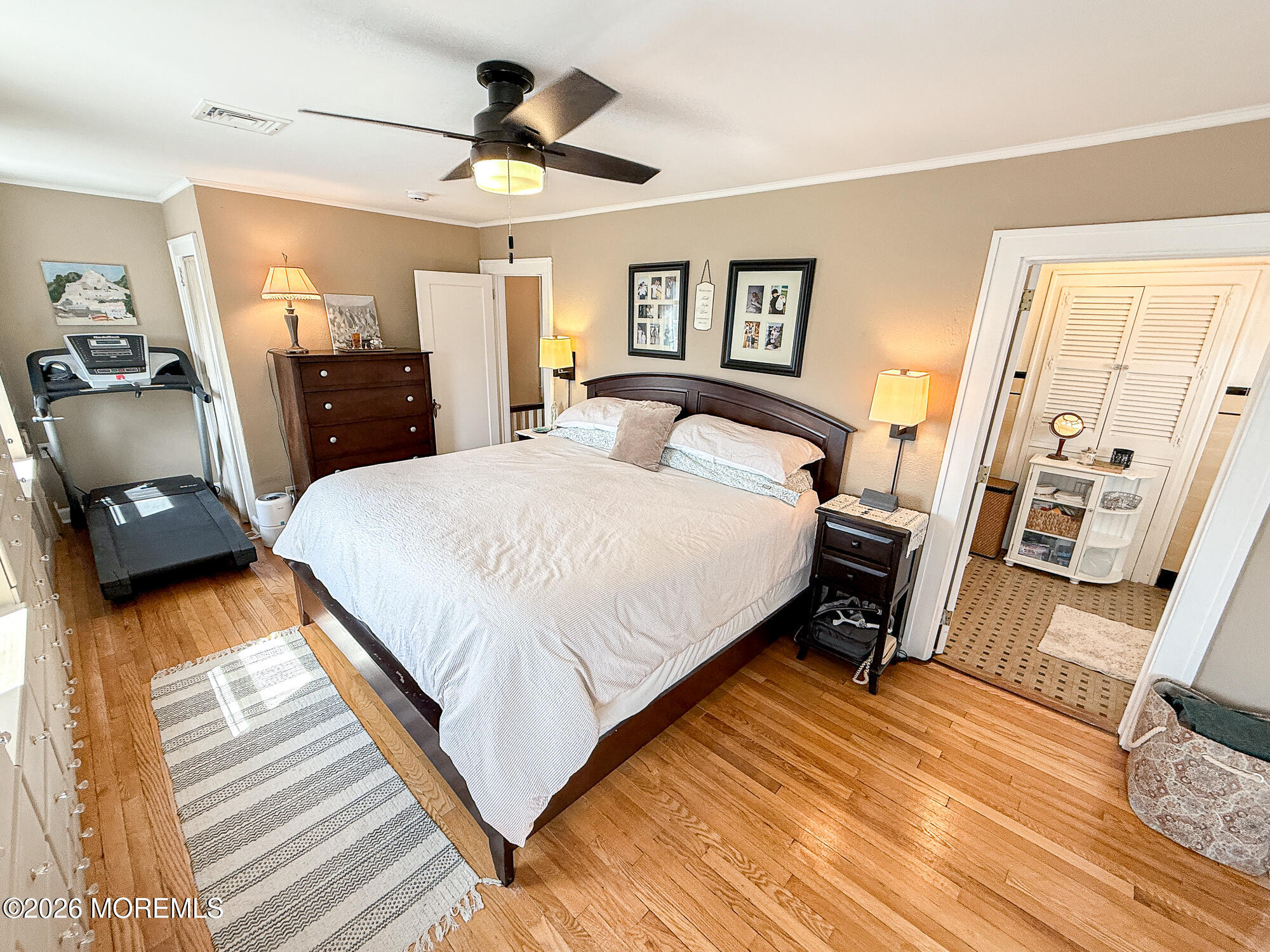 Bedroom with a large white-covered bed, hardwood floors, ceiling fan, and treadmill.