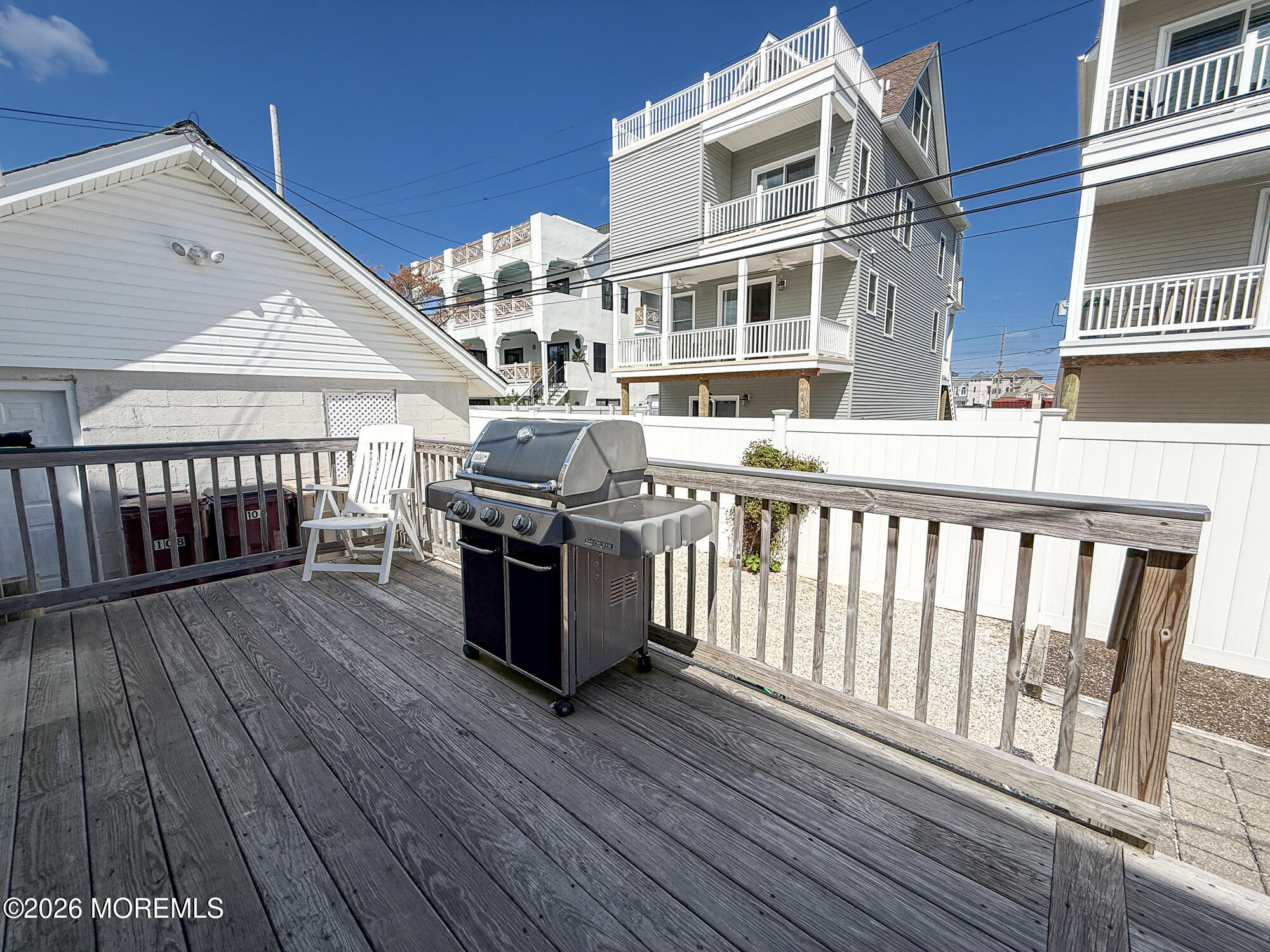 Wooden deck with a gas grill and white chair, overlooking a fenced yard and neighboring homes.