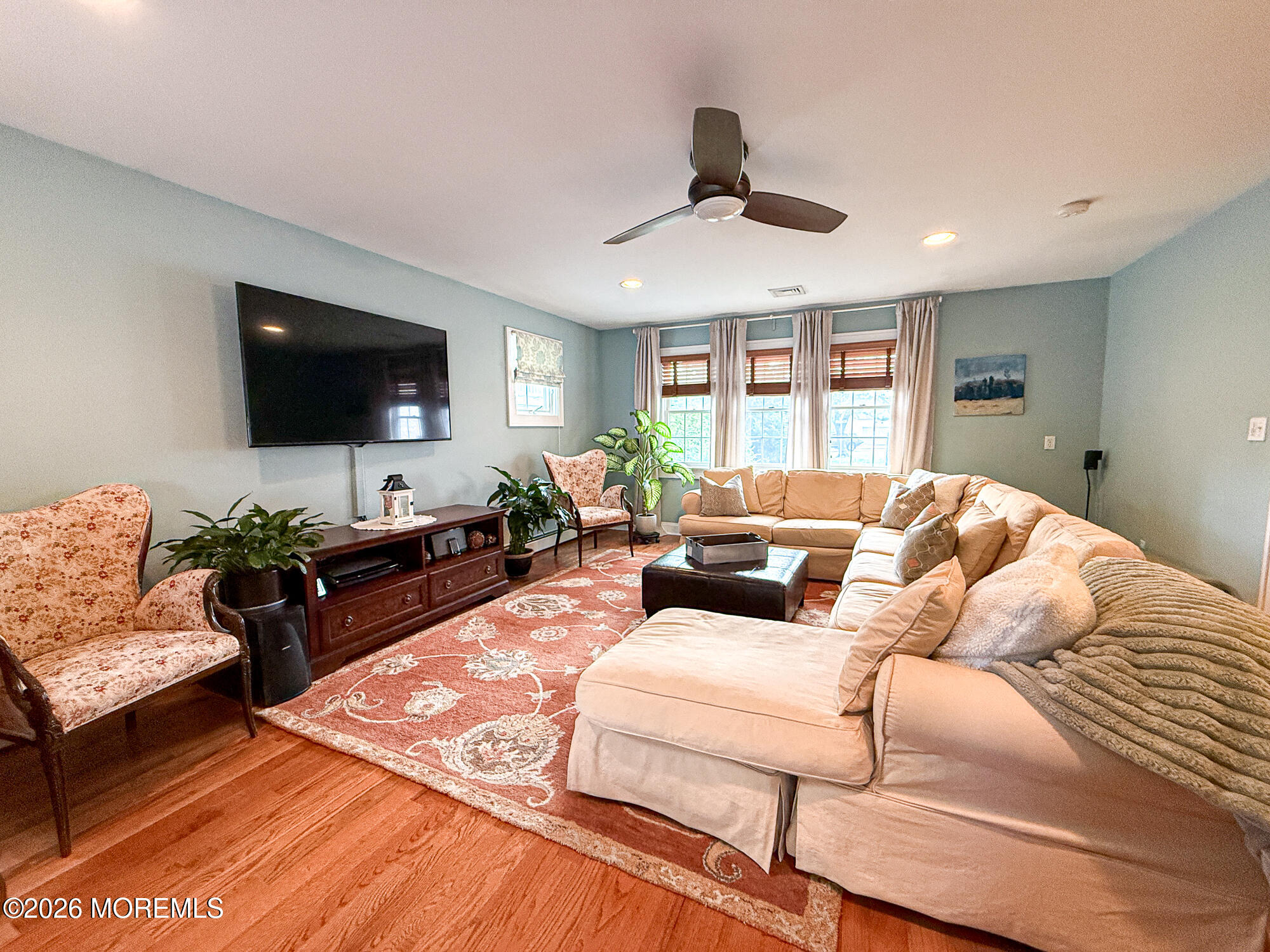 Furnished living room with a beige sectional, floral rug, wall-mounted TV, and three curtained windows.