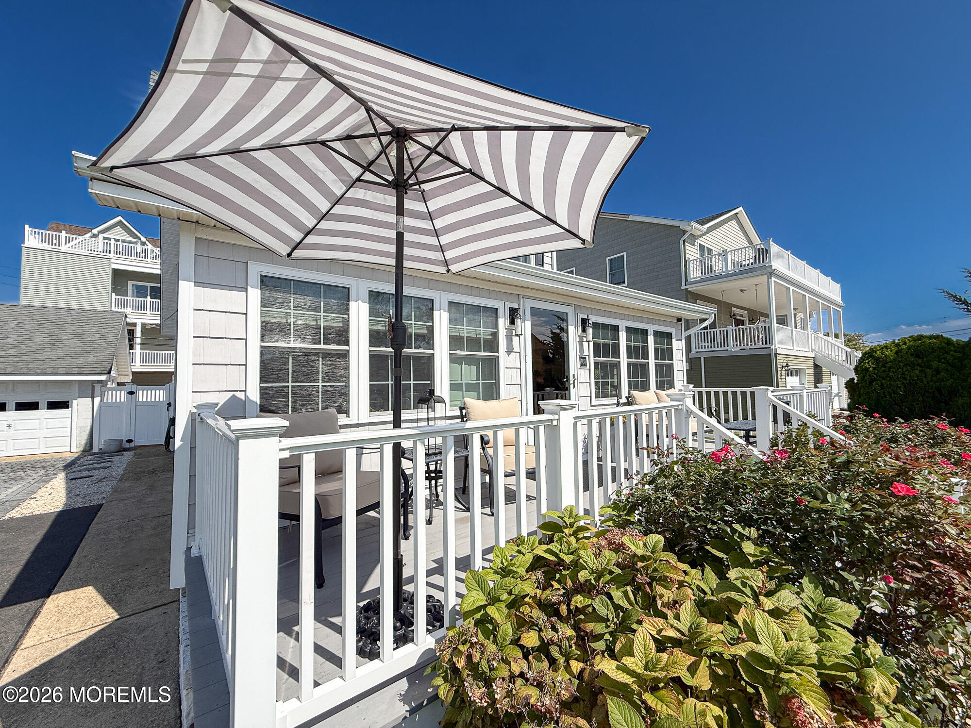 Furnished deck with striped umbrella, white railings, and flowering shrubs beside a coastal-style home.