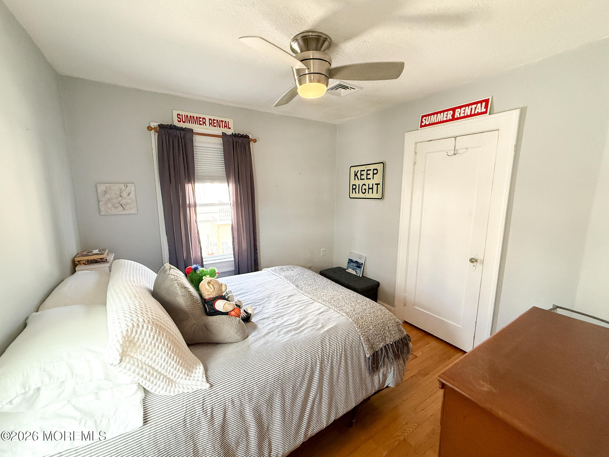 Furnished bedroom with a striped bed, purple curtains, and summer rental signs.