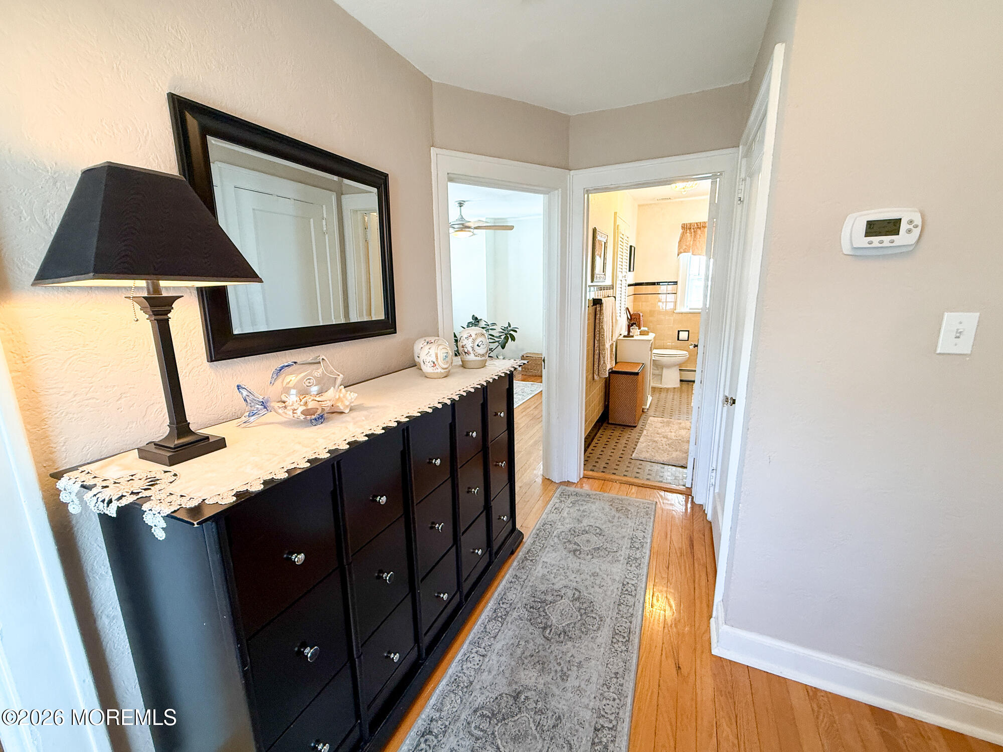 Furnished hallway with hardwood floors, a dark dresser and mirror, and open doorways to a bedroom and bathroom.