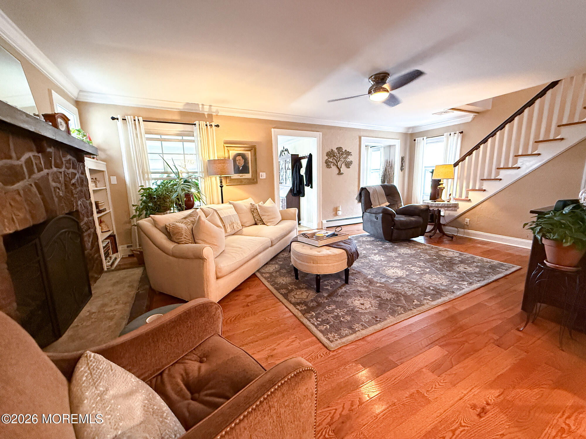 Living room with a stone fireplace, beige sofa, hardwood floors, and staircase.