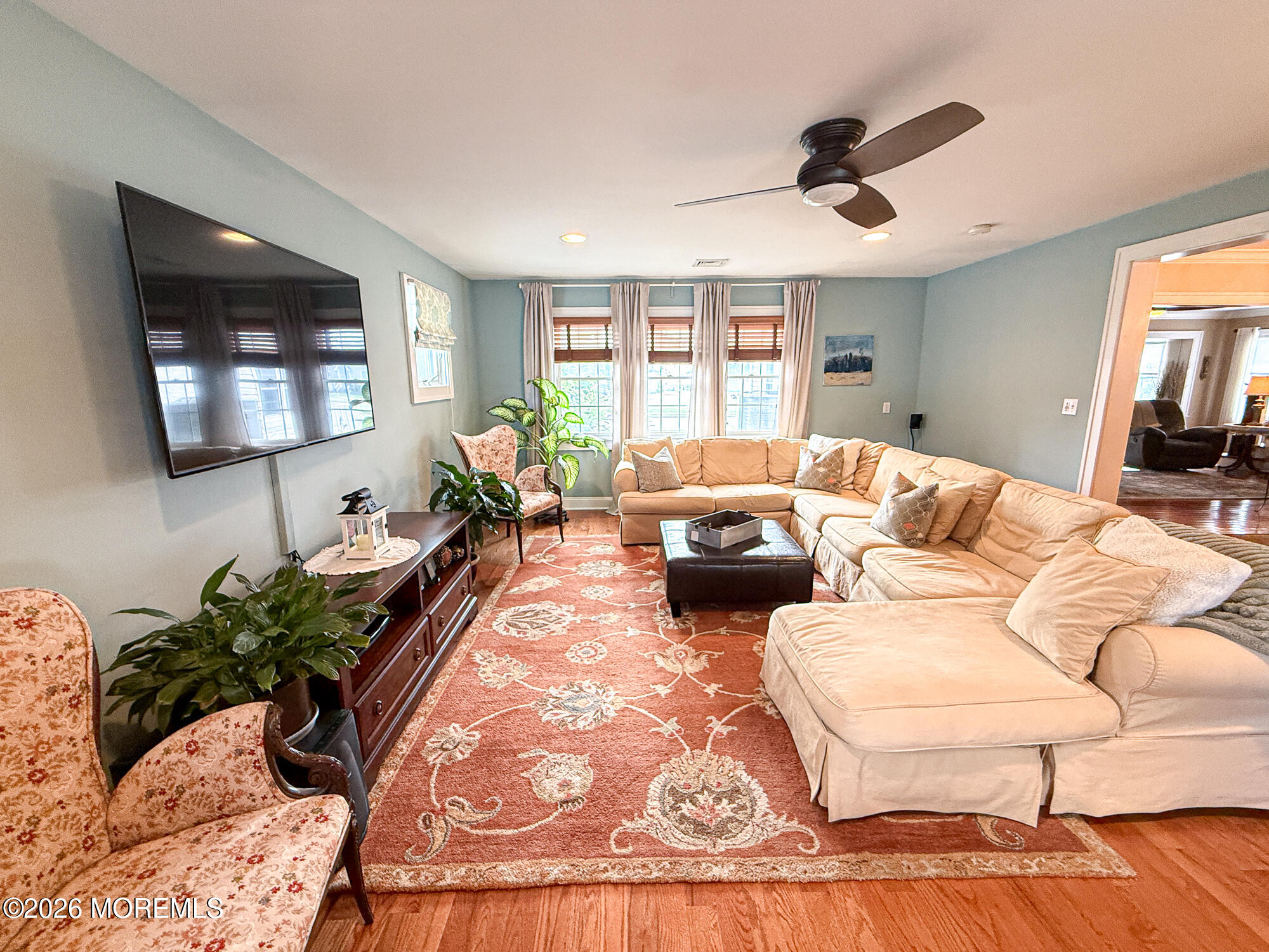 Furnished living room with a cream sectional, patterned rug, wall-mounted TV, and three curtained windows.