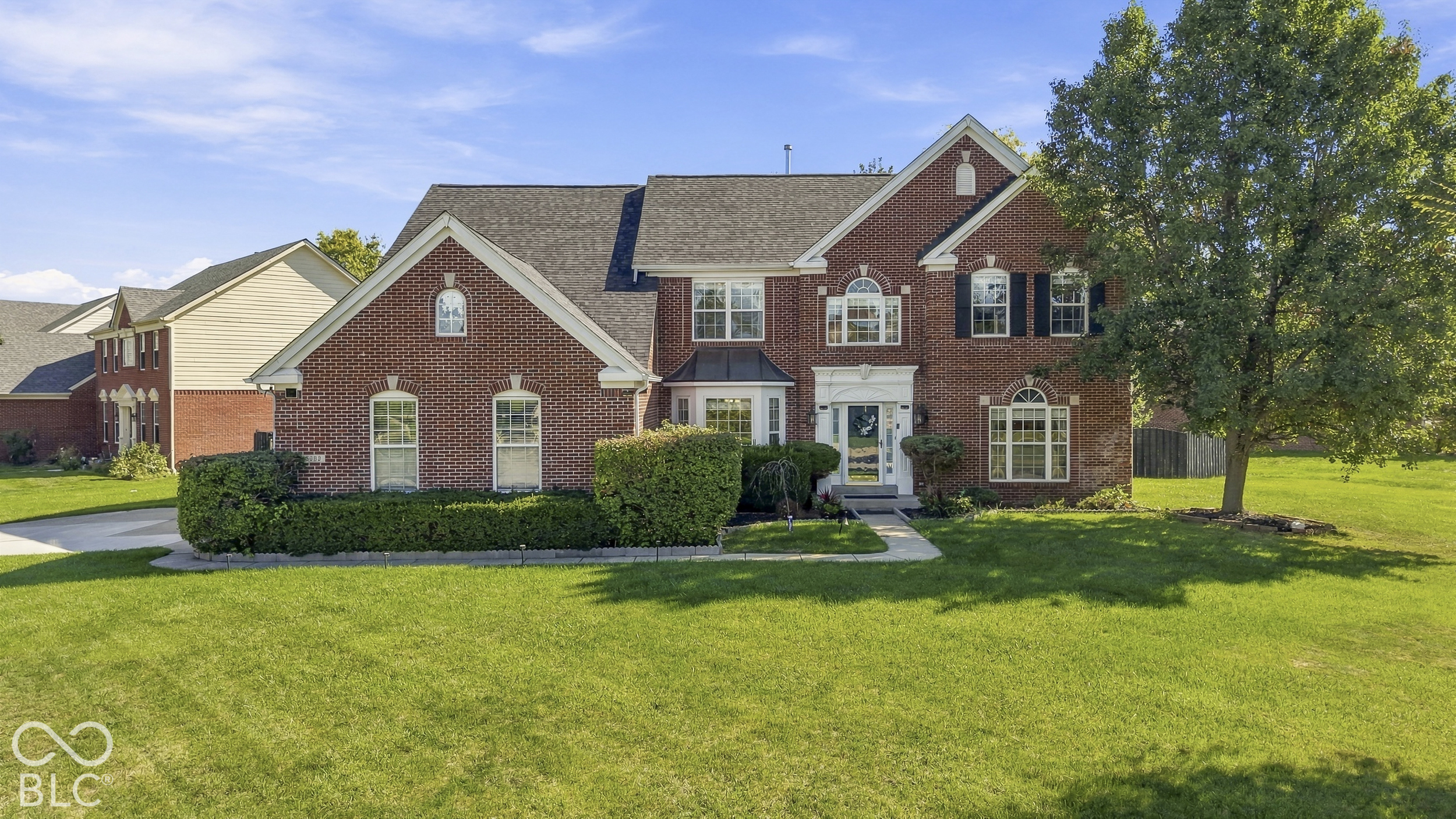 Two-story red brick house with white trim, landscaped shrubs, and a broad green lawn.