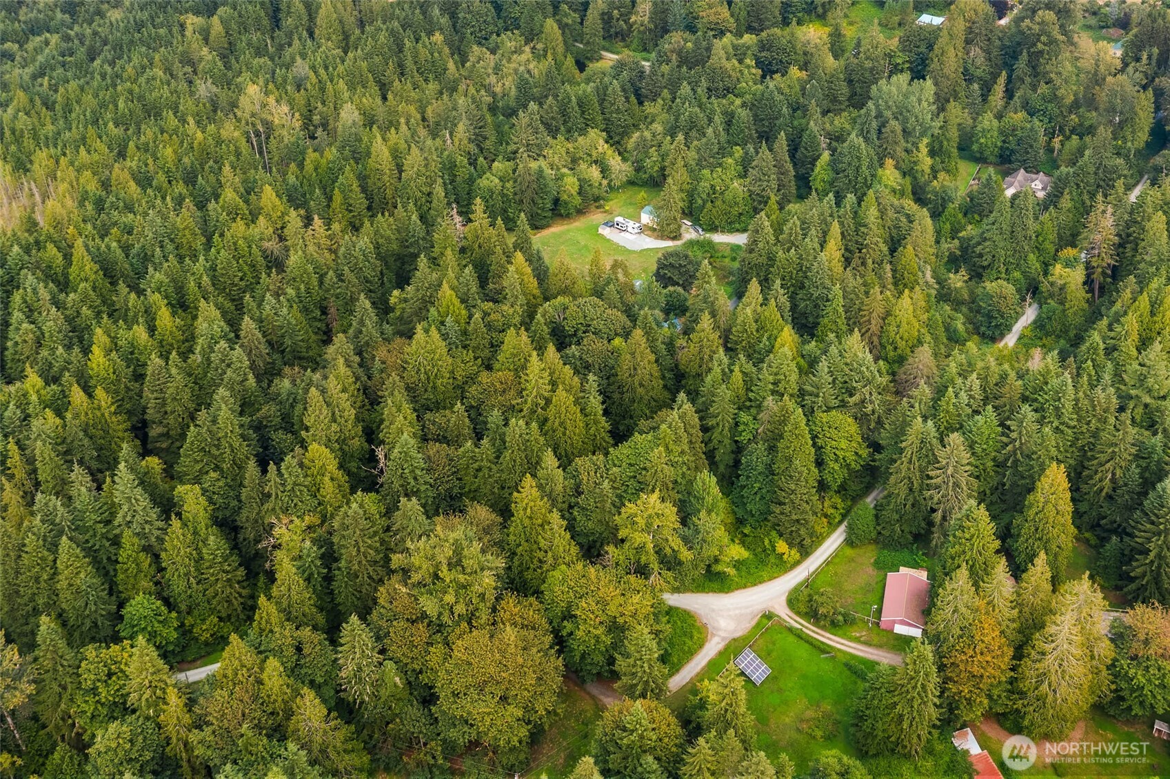 Aerial view of a forested property with a clearing, RV, scattered buildings, and winding access roads.