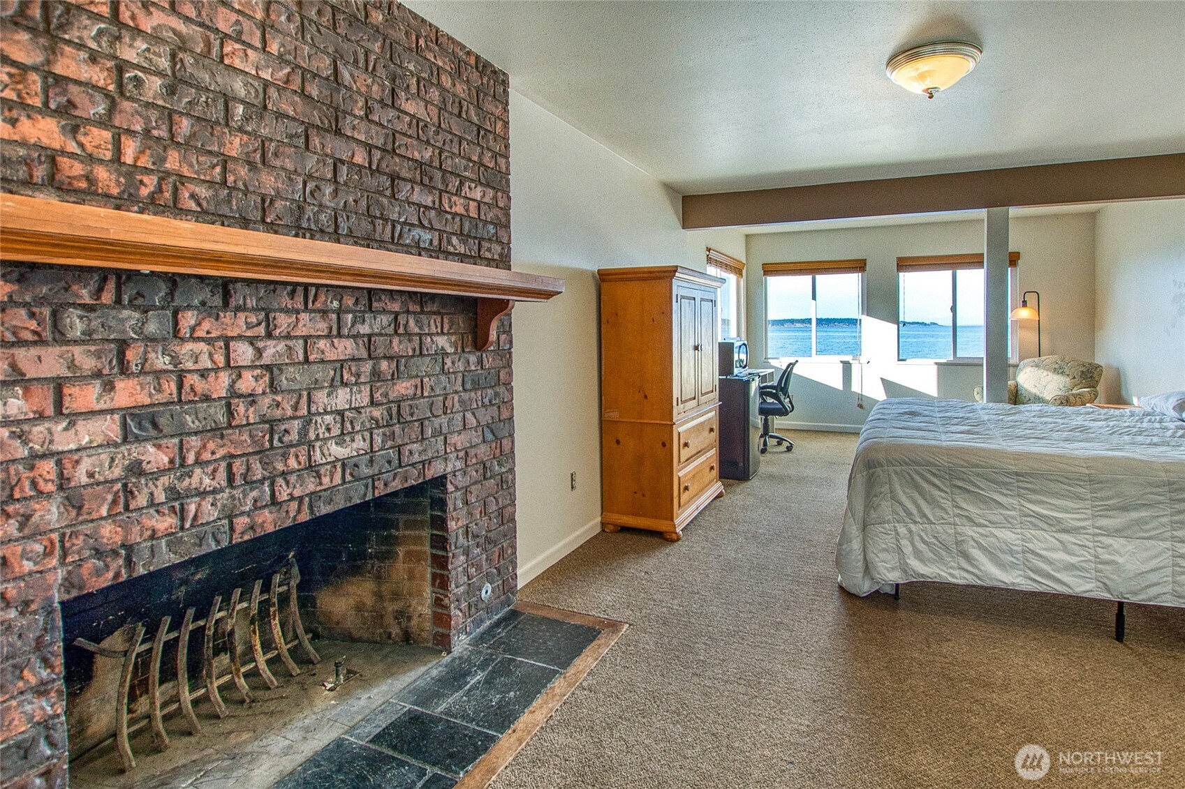 Bedroom with a brick fireplace, bed, and windows overlooking the water.