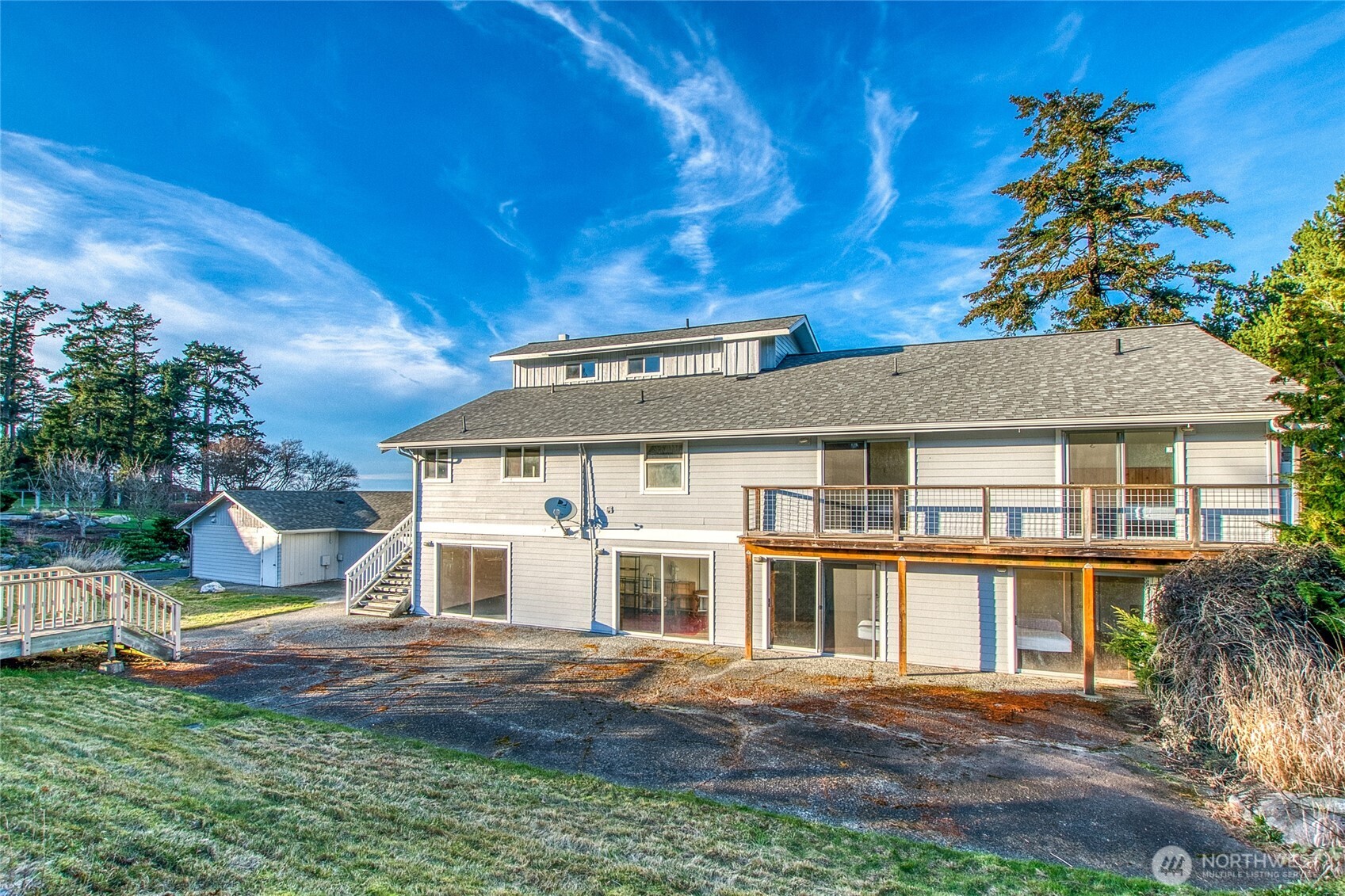 Two-story gray home with a long deck, patio doors, detached outbuilding, and a glimpse of water.