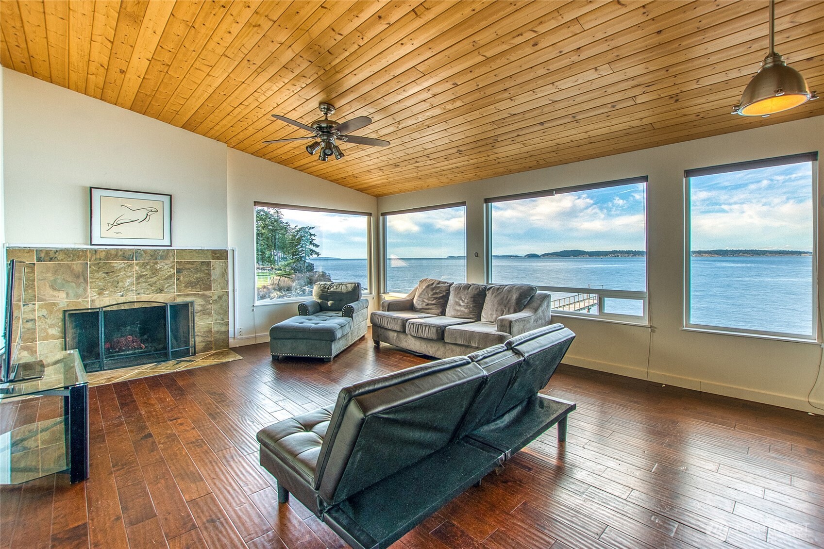 Living room with a tiled fireplace, wood ceiling, and wide water views through large windows.