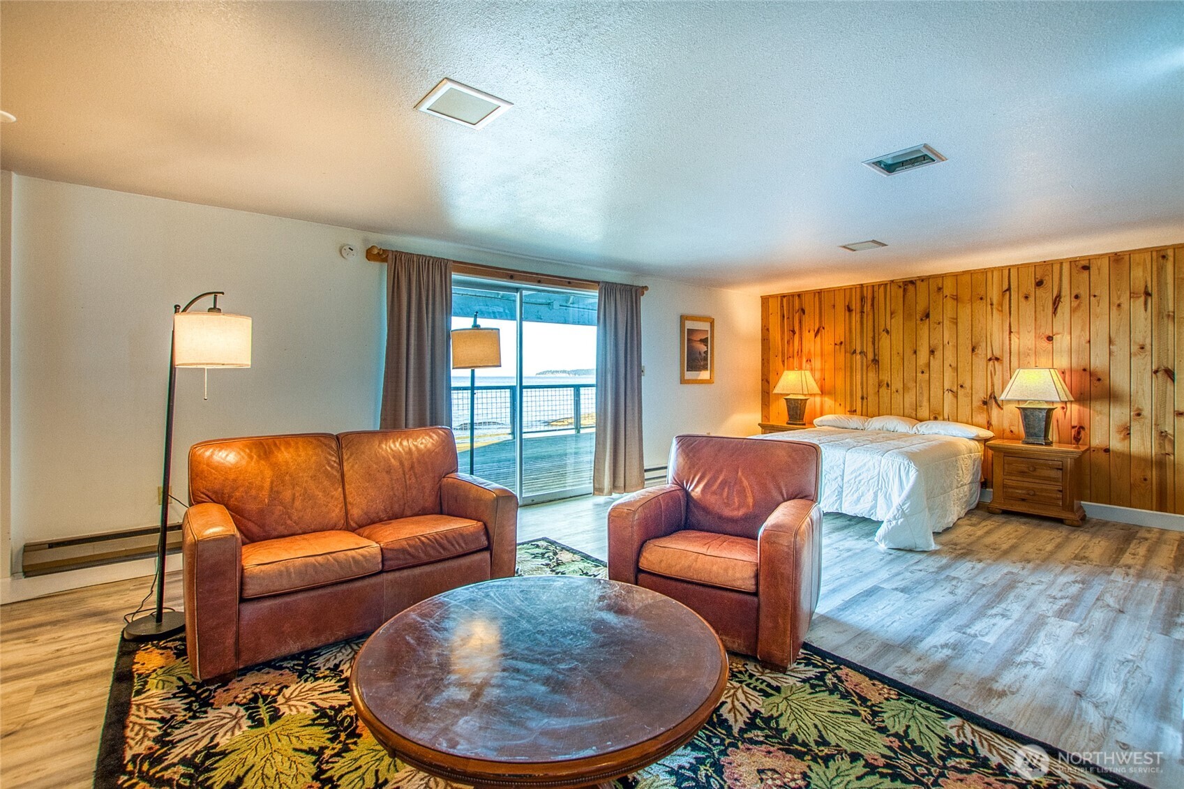 Furnished bedroom and sitting area with leather seating, pine-paneled wall, and water visible beyond the sliding doors.