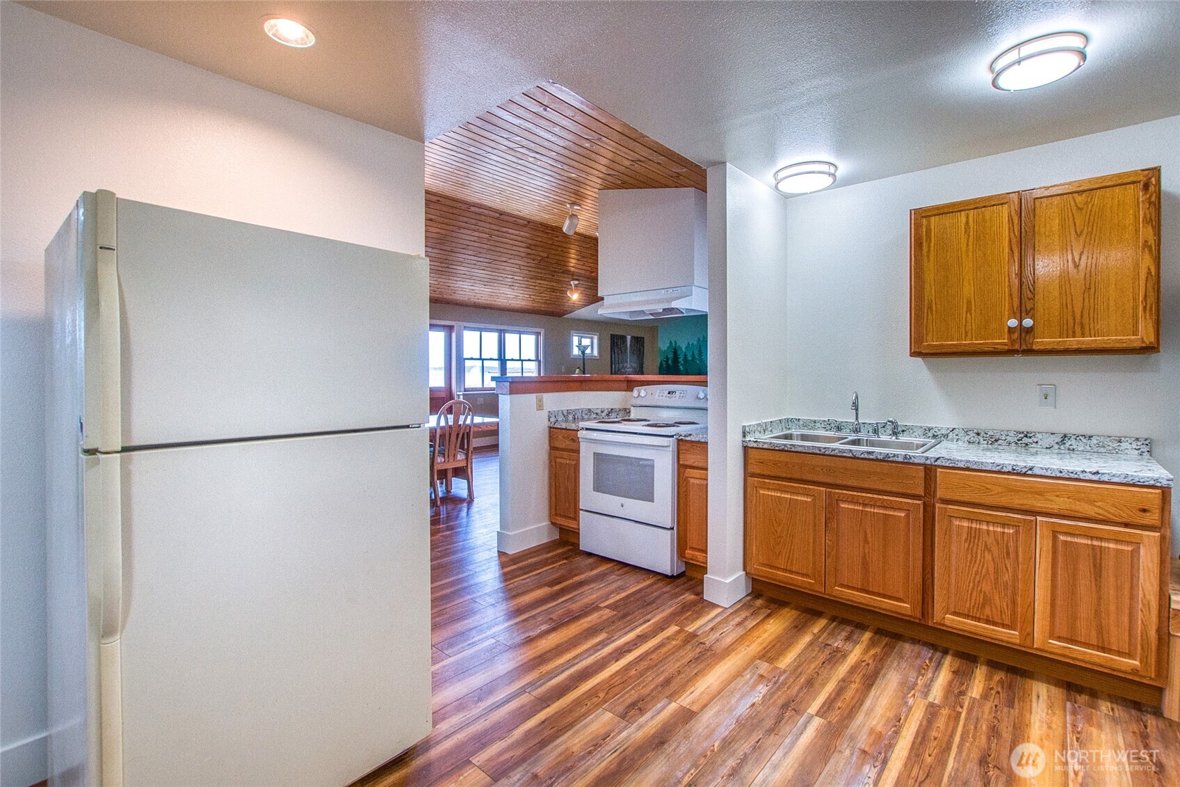 Kitchen with wood cabinetry and flooring, white appliances, and an adjoining dining area beneath a vaulted wood ceiling.