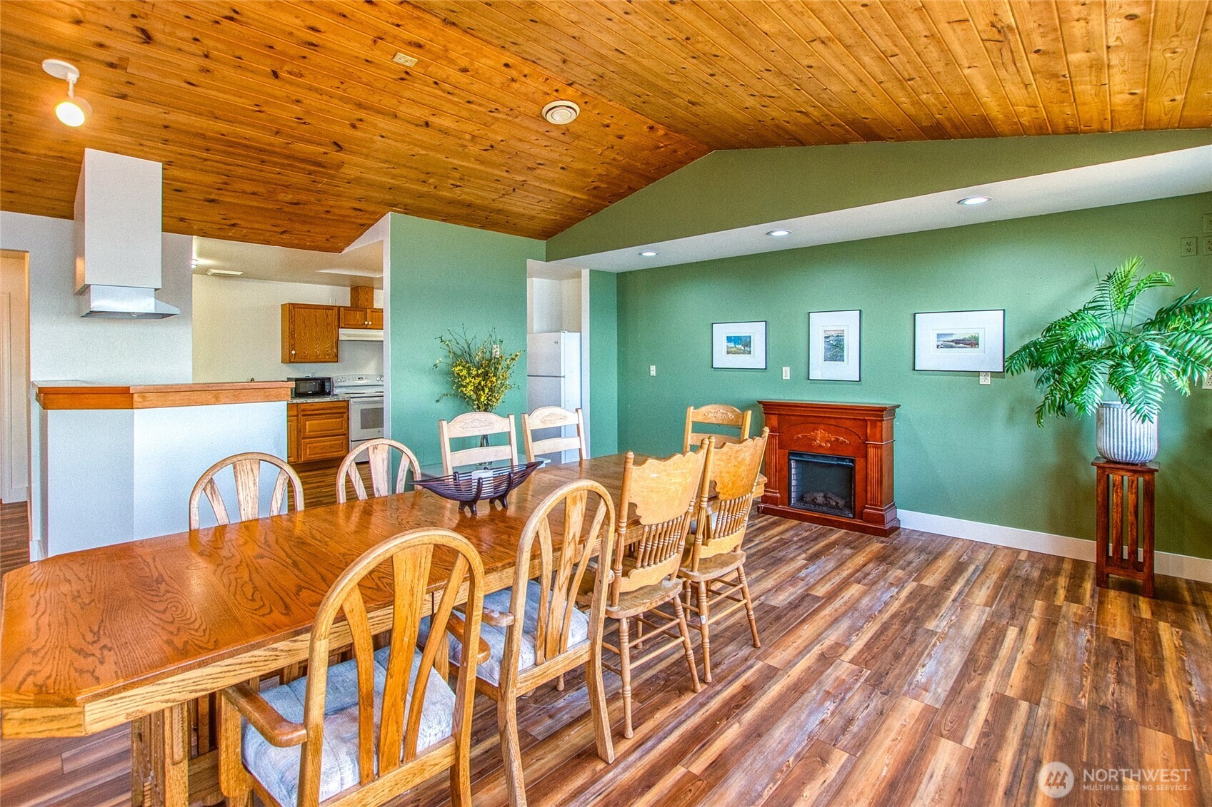 Dining area with a long wooden table beneath a wood-plank ceiling, open to the kitchen.