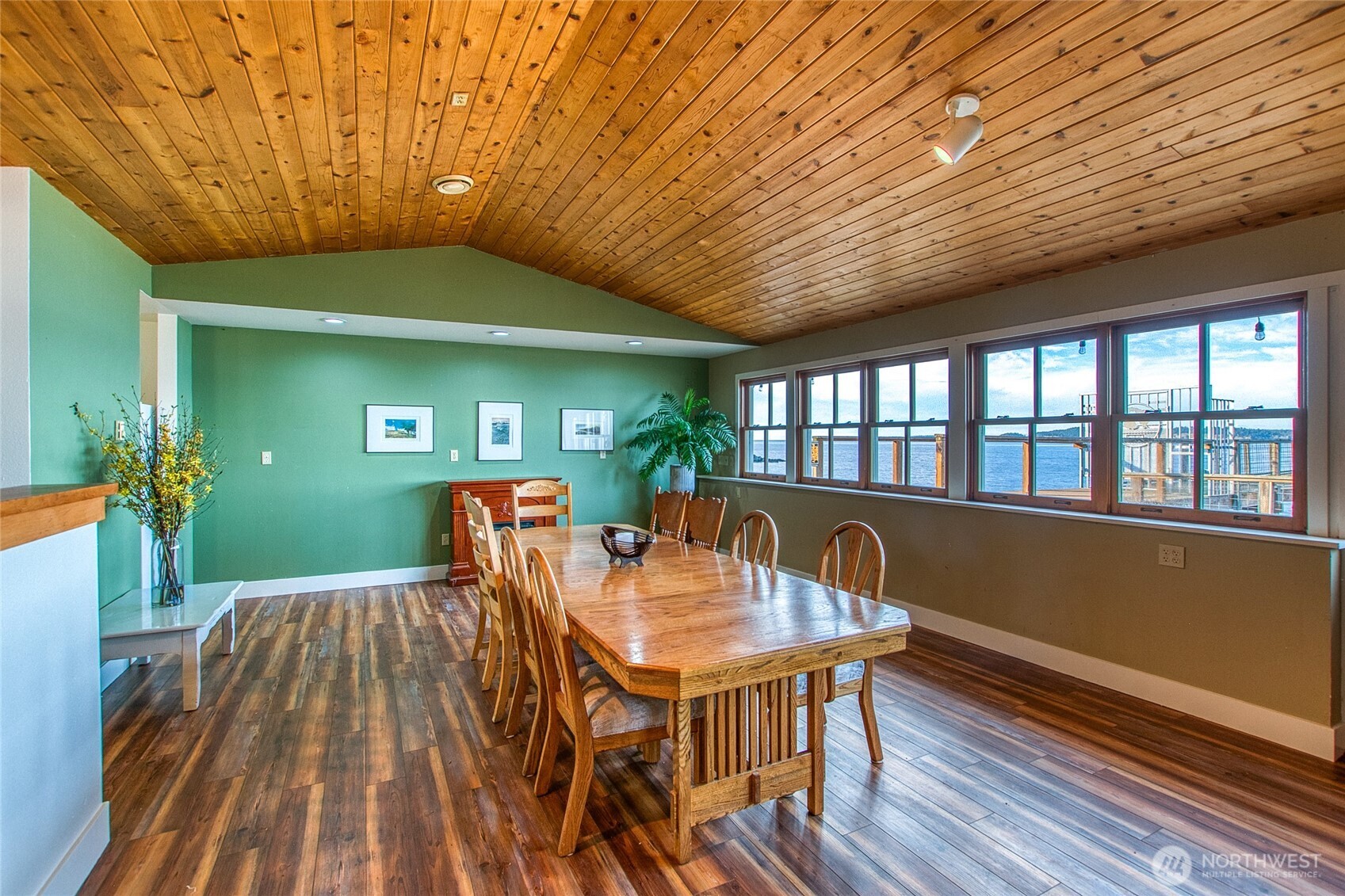 Dining room with a long wooden table, vaulted wood ceiling, and windows overlooking the water.