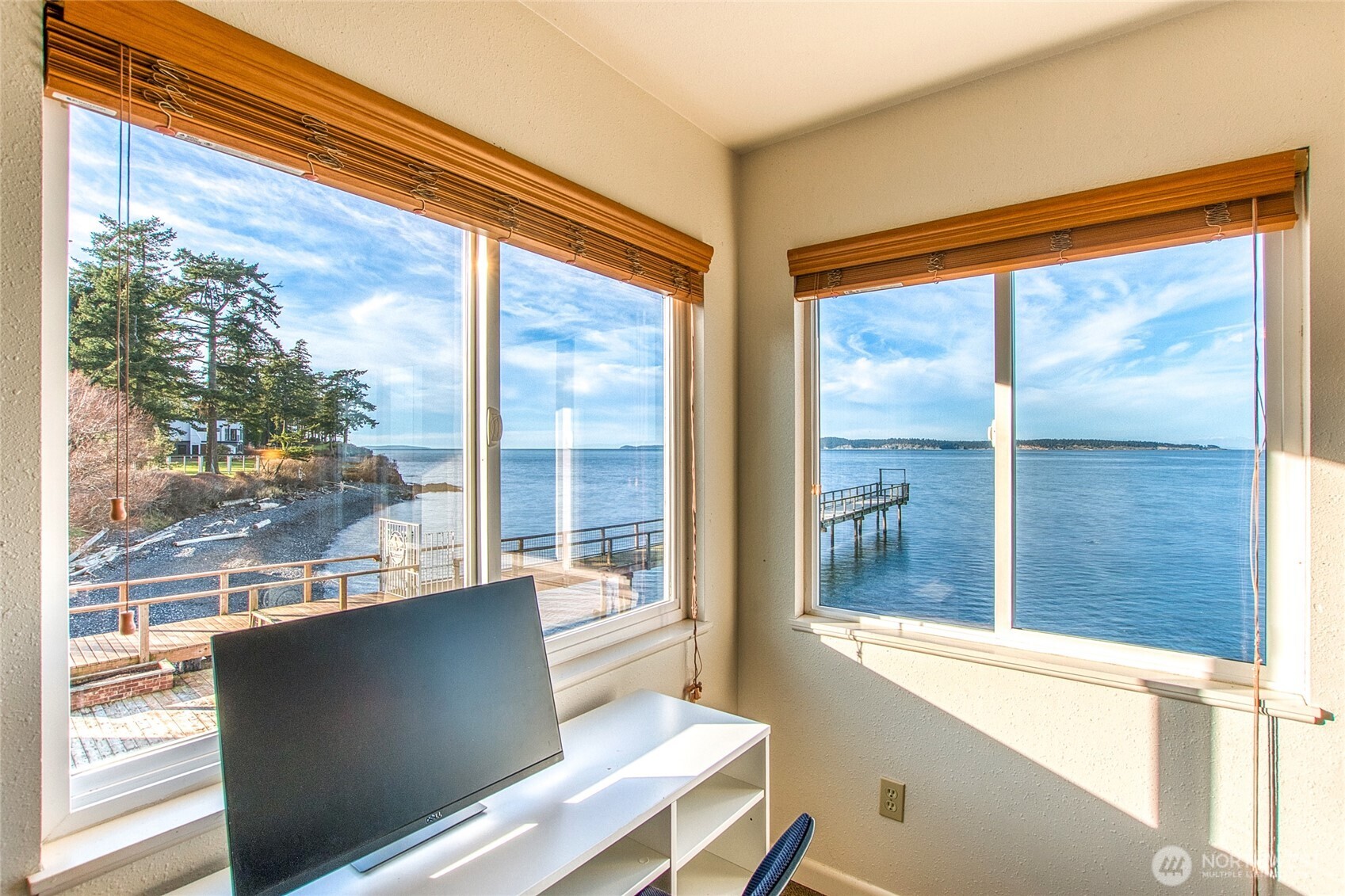 Waterfront room with a desk and monitor, framed by two bay-facing windows and a view of a pier.