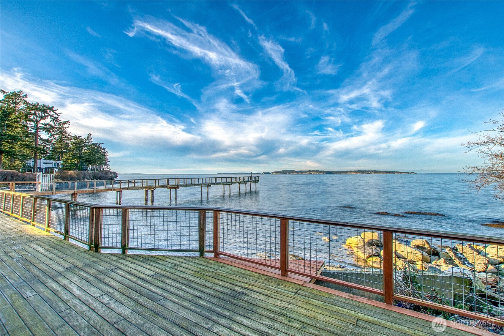 Waterfront deck overlooking a long pier, rocky shore, calm water, and distant islands.
