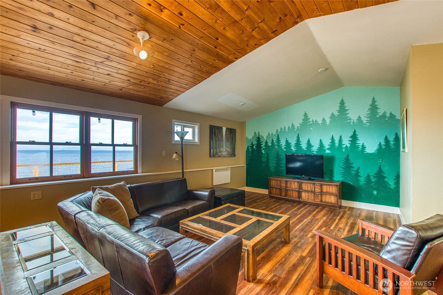 Living room with leather seating, a forest mural, and water views through large windows.