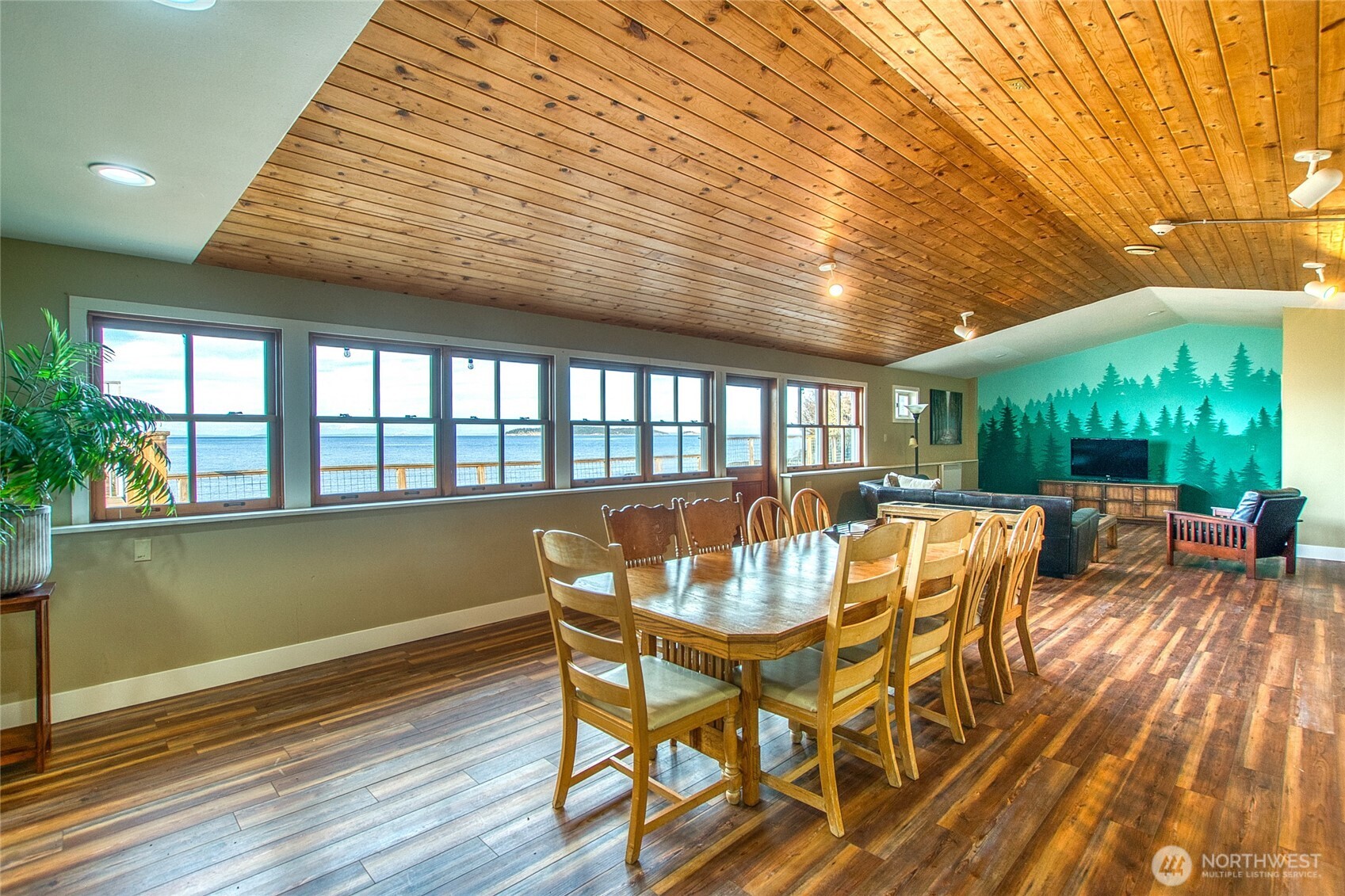 Dining and living room with a wood-plank ceiling, teal forest mural, and water views through a row of windows.