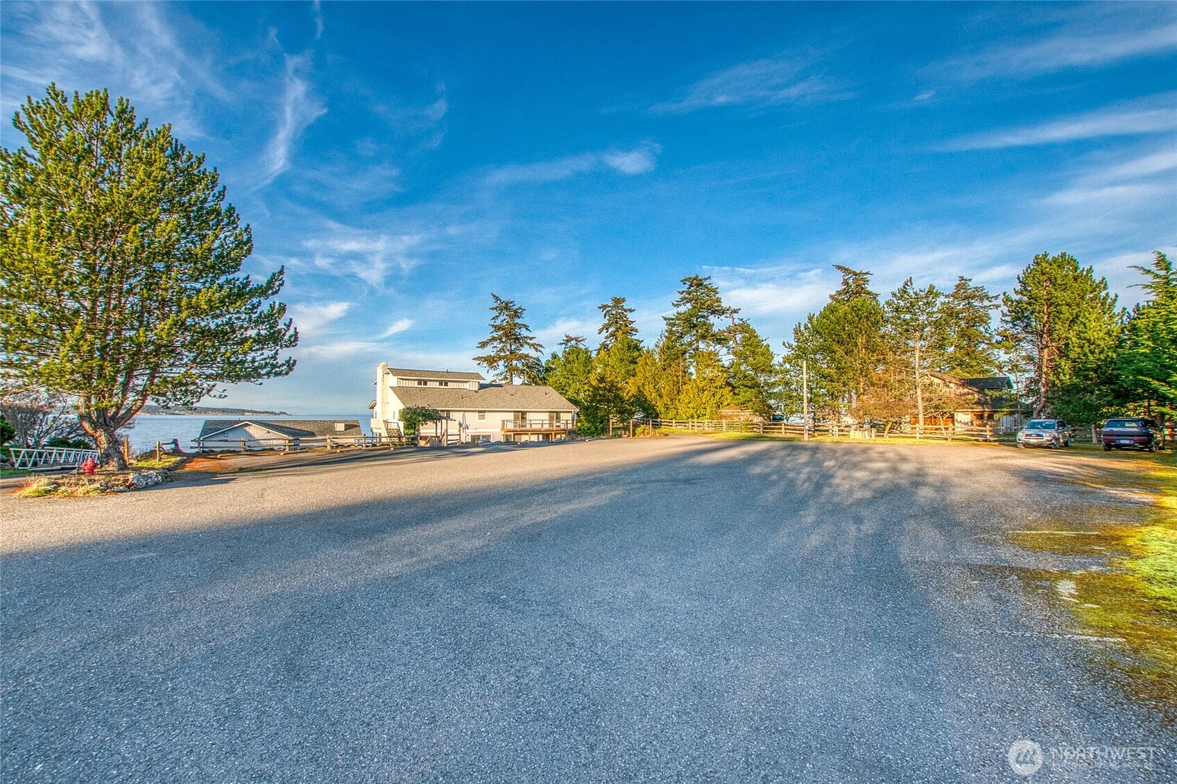 Waterfront property with a broad paved parking area, evergreen trees, and shoreline views.
