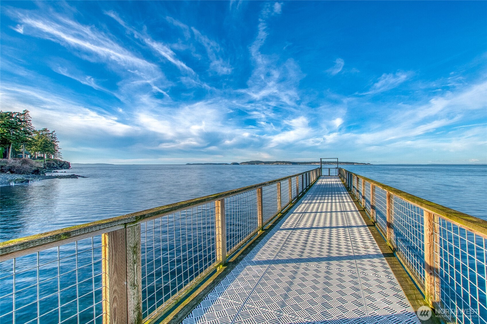 A metal-grate pier extends over calm water toward a distant wooded island.