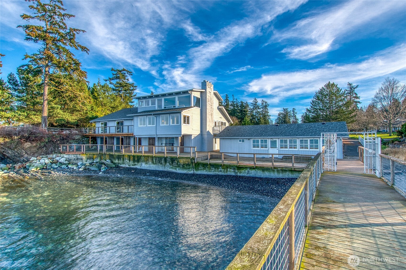 Waterfront house with a stone chimney, deck, and boardwalk along a rocky shore.