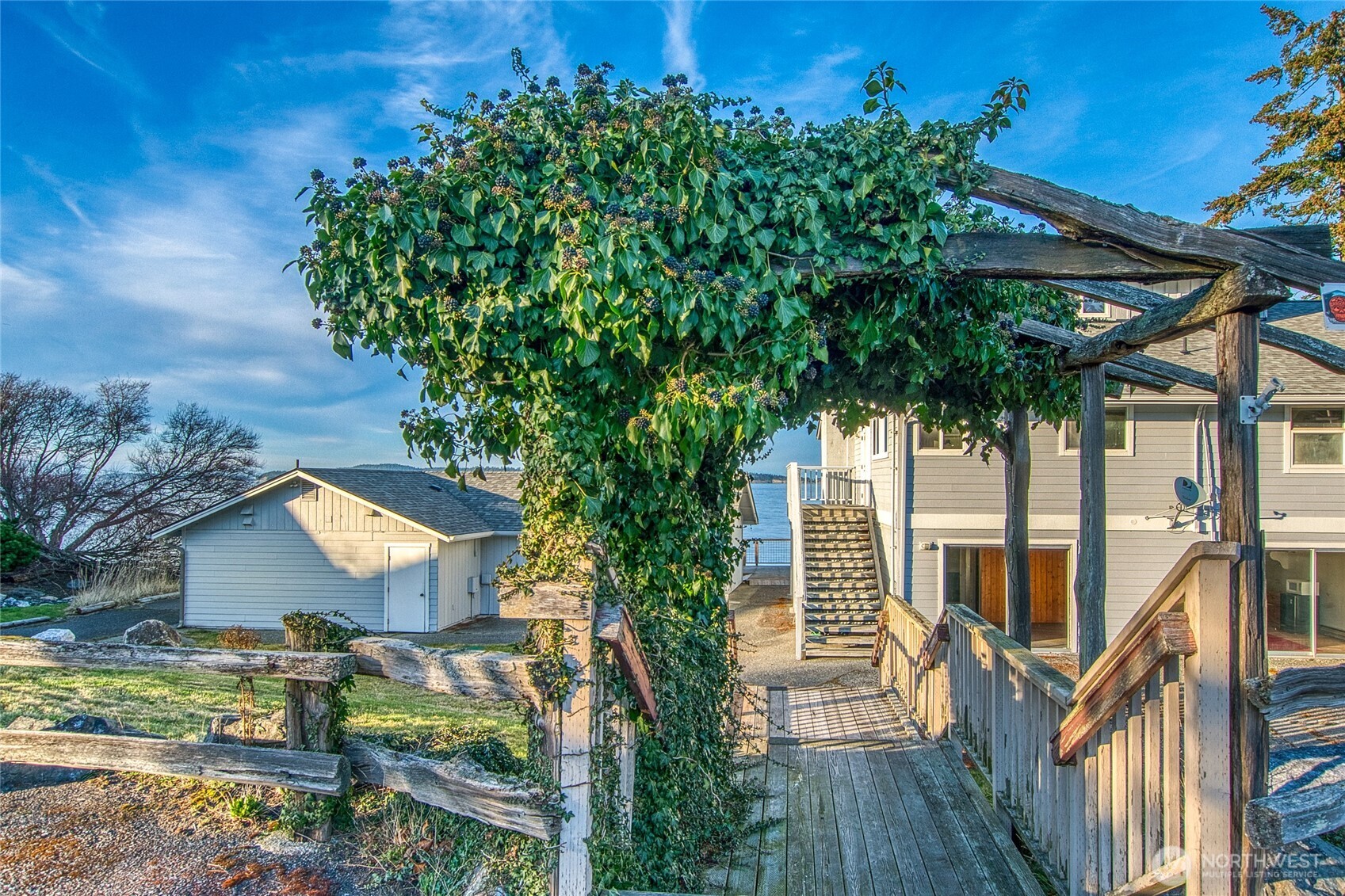 Waterfront home with an ivy-covered pergola over a wooden walkway and the bay beyond.