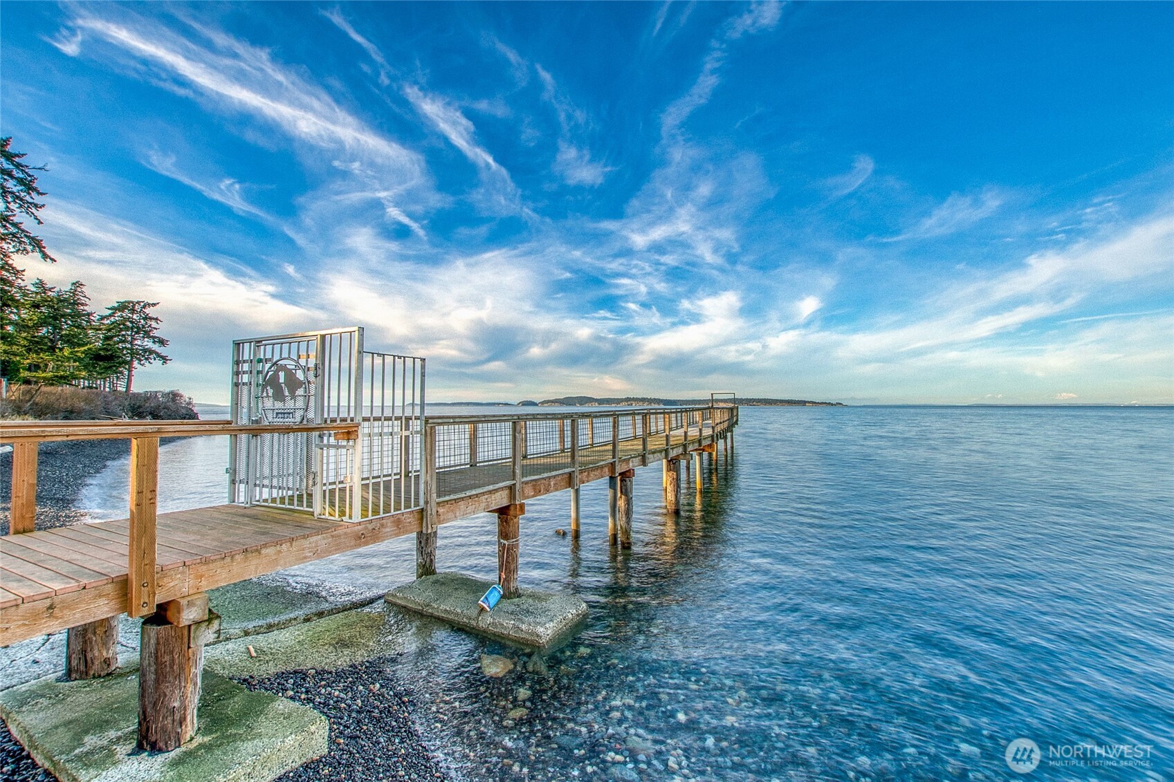 Wooden pier extending over calm water from a rocky shoreline beneath a blue sky.
