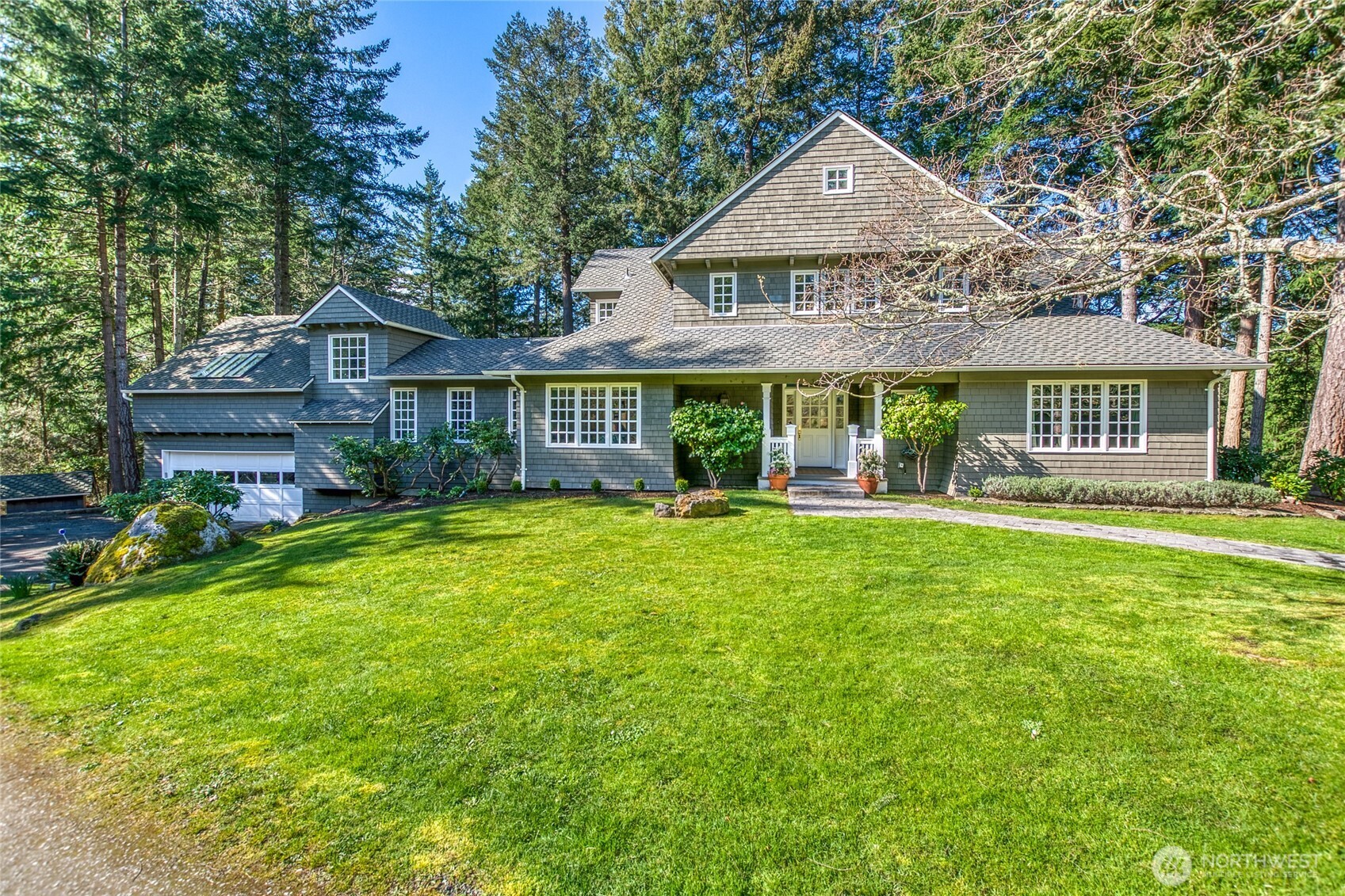 Gray shingle-sided house with multiple gables, an attached garage, and a broad lawn among tall evergreen trees.