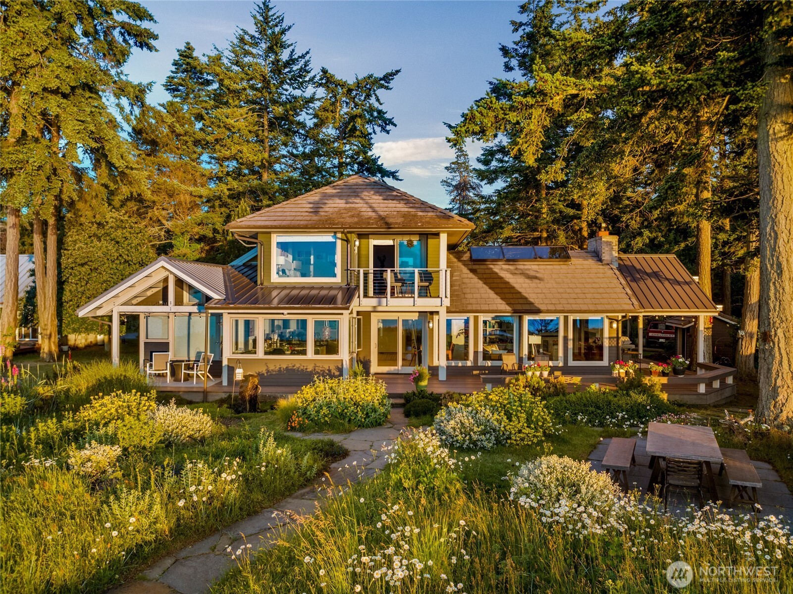 Two-story home with a broad deck, mature evergreens, and a flowering garden in golden-hour light.