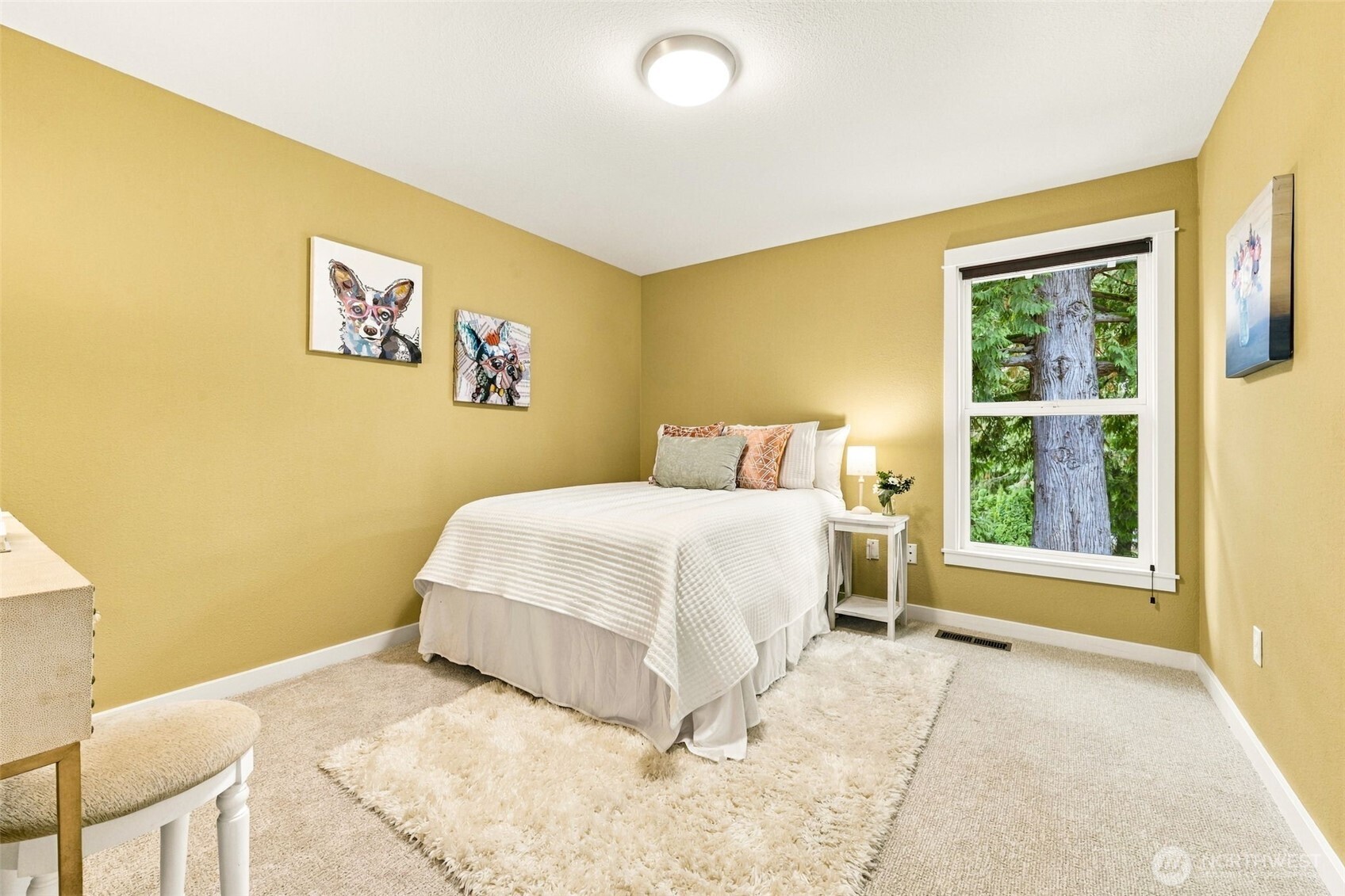 Furnished bedroom with mustard-yellow walls, a white bed, layered cream rugs, and a large window overlooking a tree.