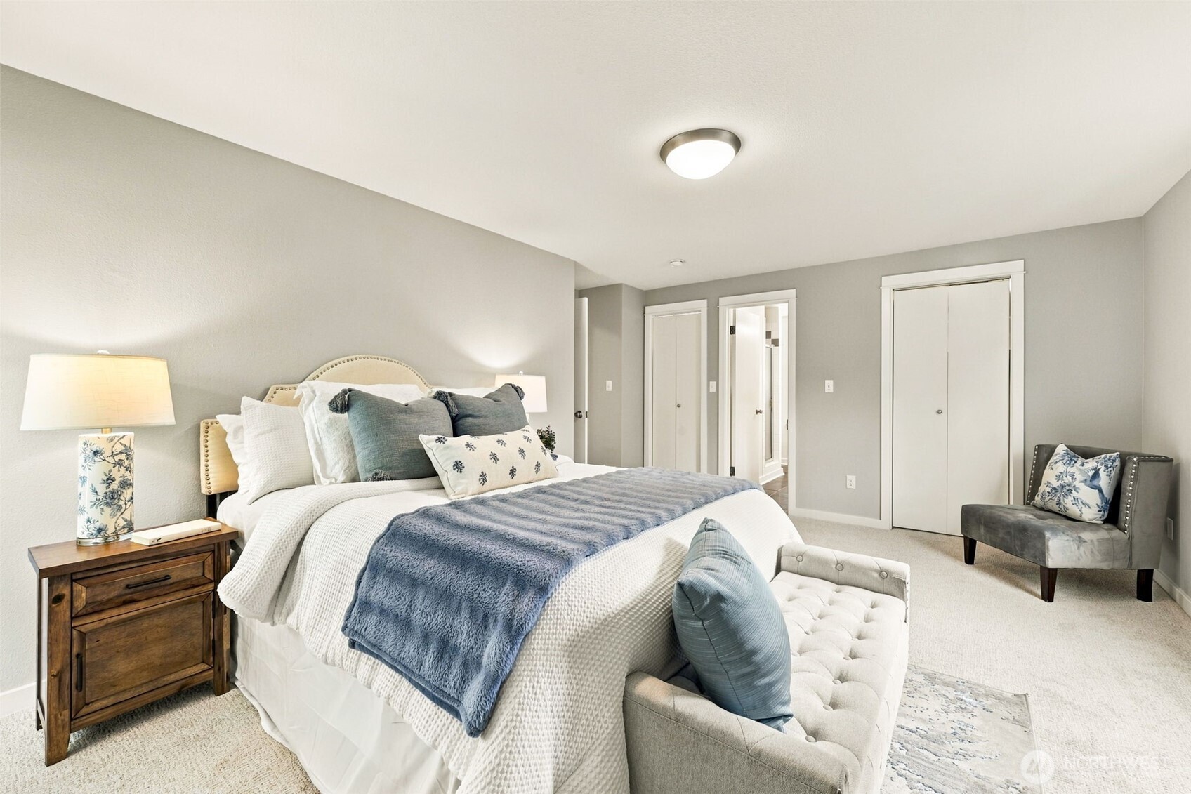 Staged bedroom with a blue throw on the bed, tufted bench, wooden nightstand, and gray upholstered chair.