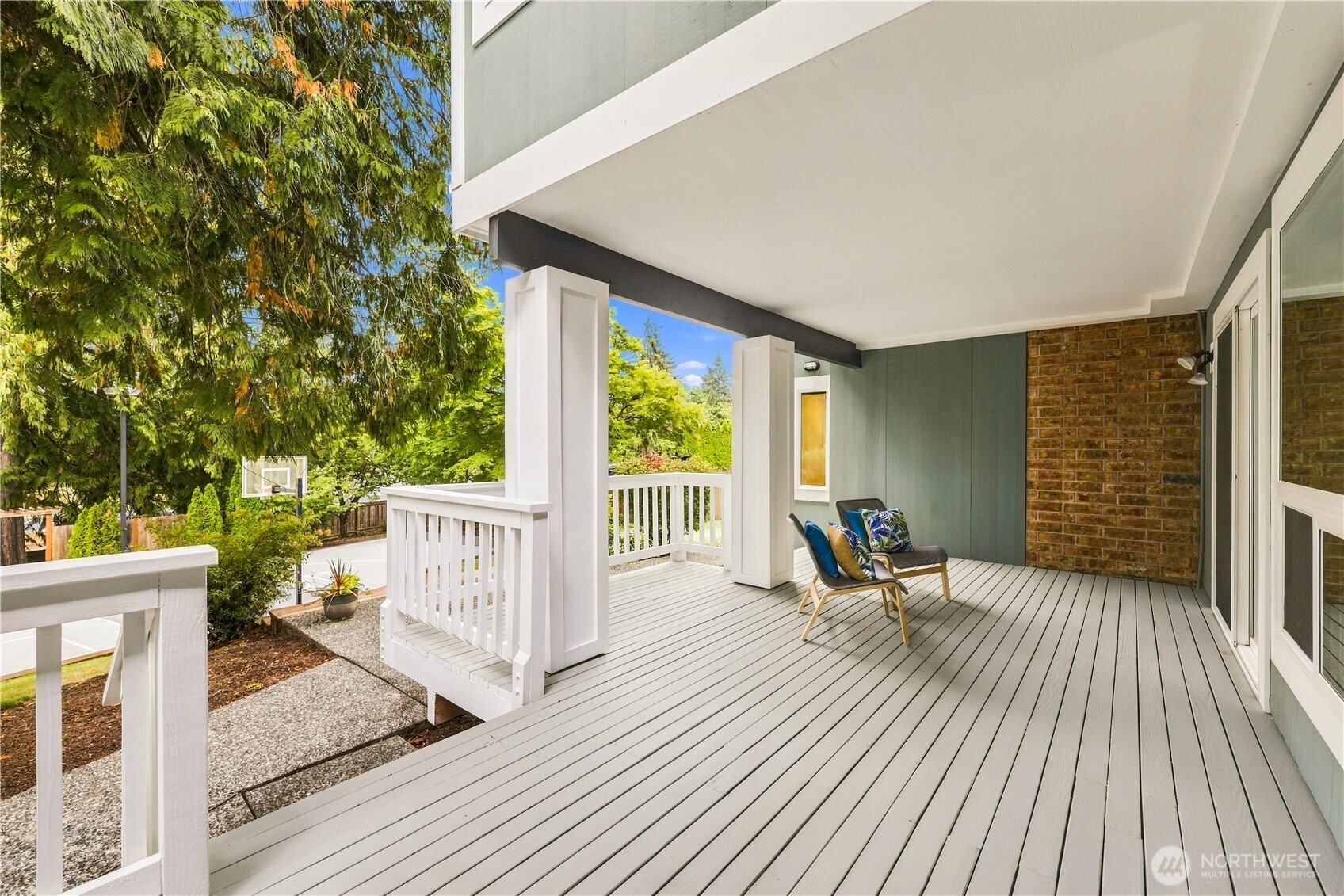 Covered deck with white railings, brick accent wall, and colorful seating overlooking leafy trees.
