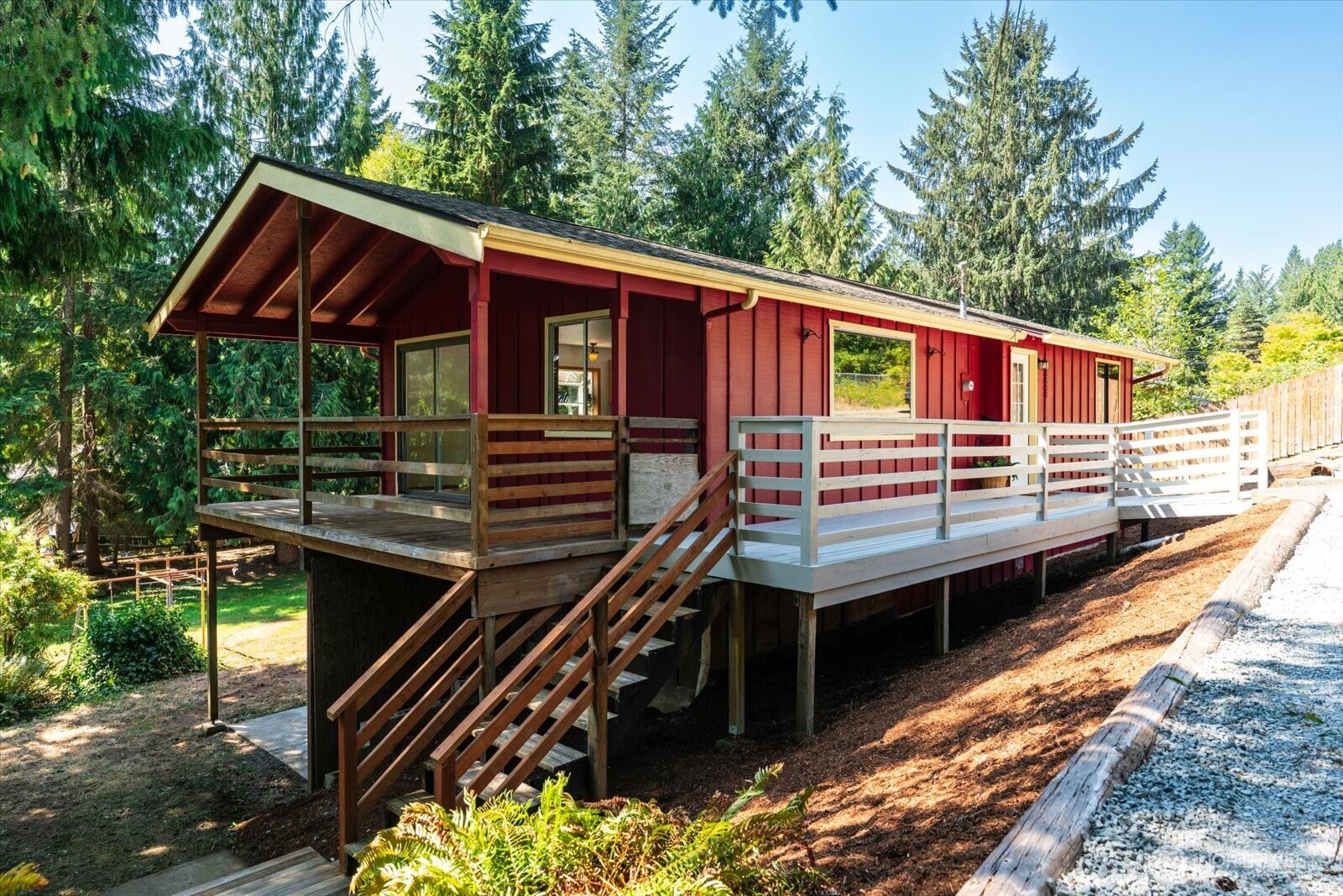Red-sided home with an elevated wraparound deck and stairs, surrounded by evergreen trees.