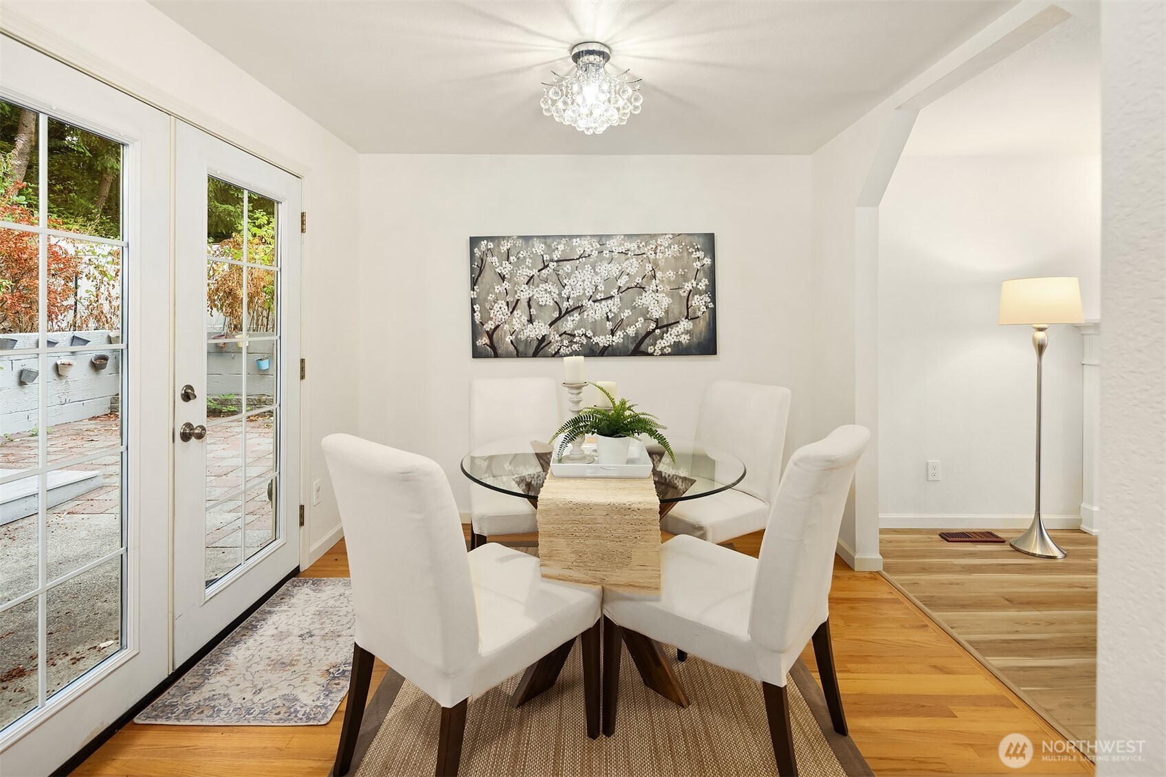 Dining room with a glass table, white chairs, crystal chandelier, and French doors opening to a patio.