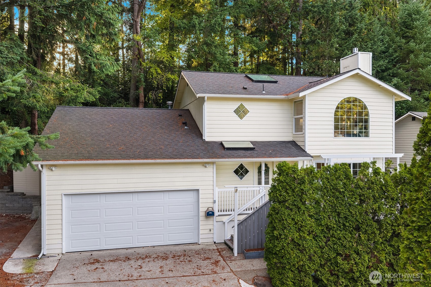 Cream two-story home with an attached double garage, arched window, and evergreen landscaping.