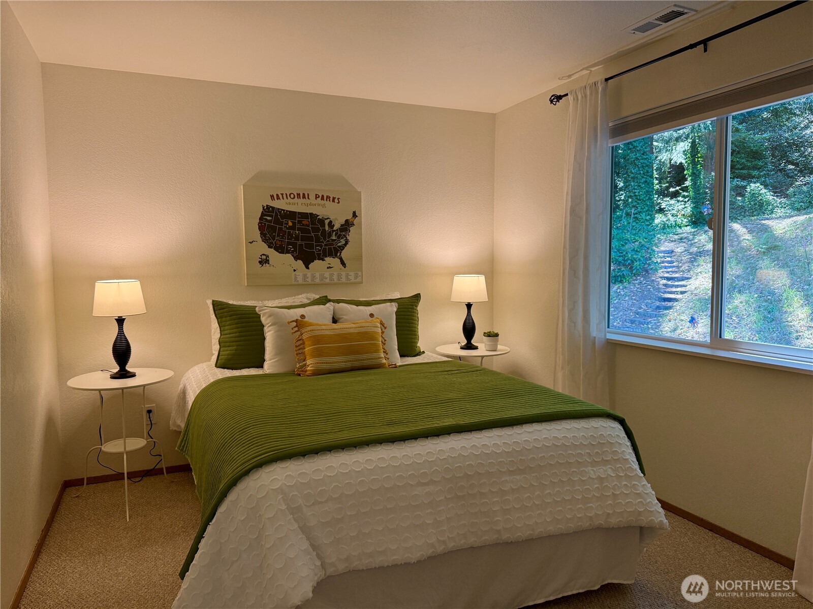 Staged bedroom with a green-covered bed, matching lamps, and a window overlooking leafy steps.
