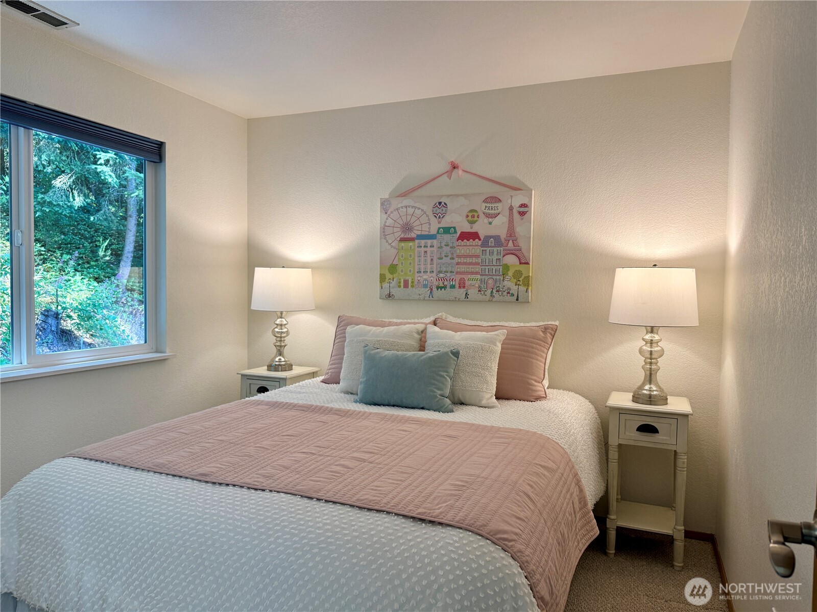 Staged bedroom with blush and pale blue bedding, matching bedside lamps, and a window overlooking greenery.