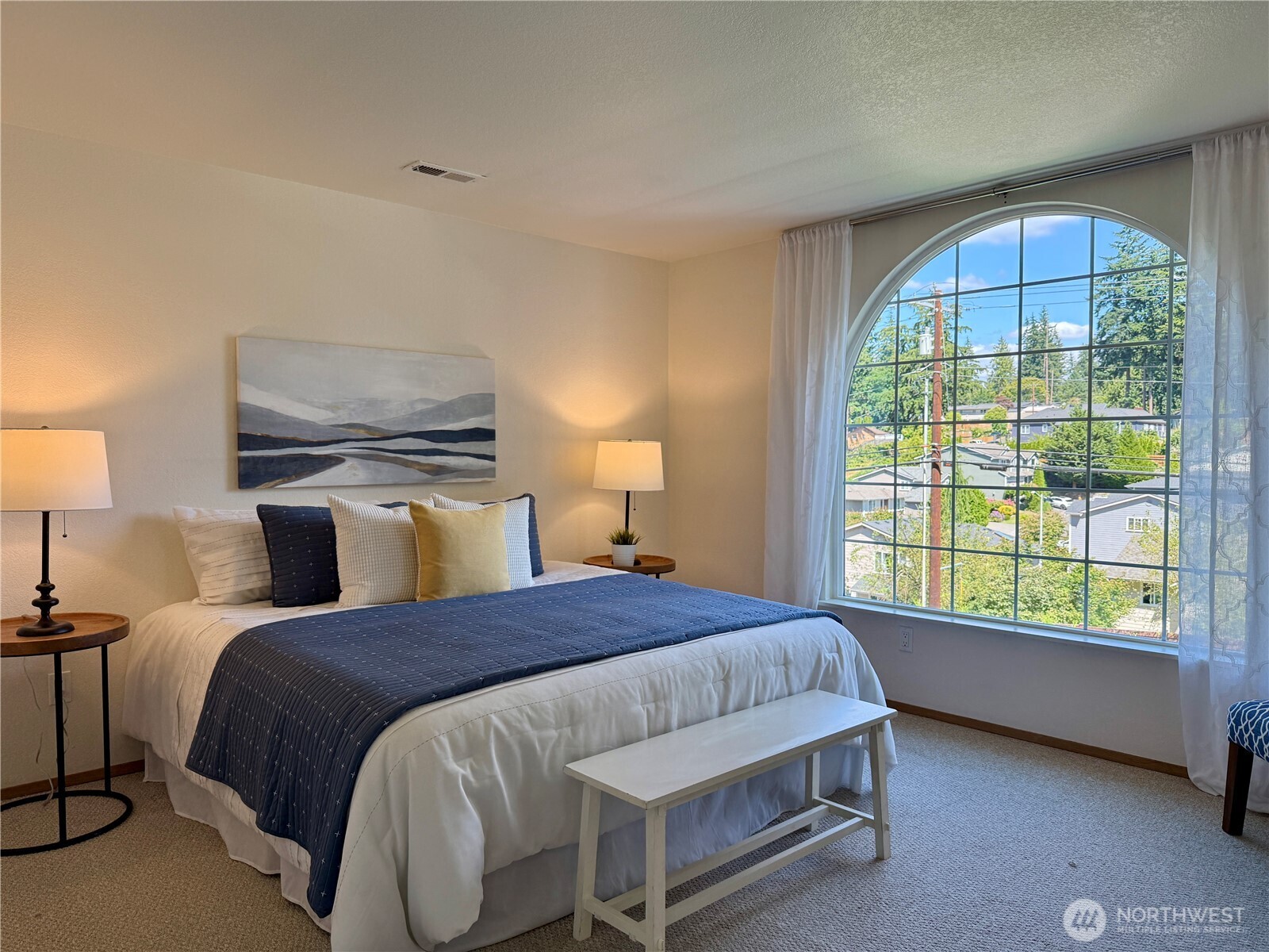 Staged bedroom with a navy-and-white bed and a large arched window overlooking homes and trees.