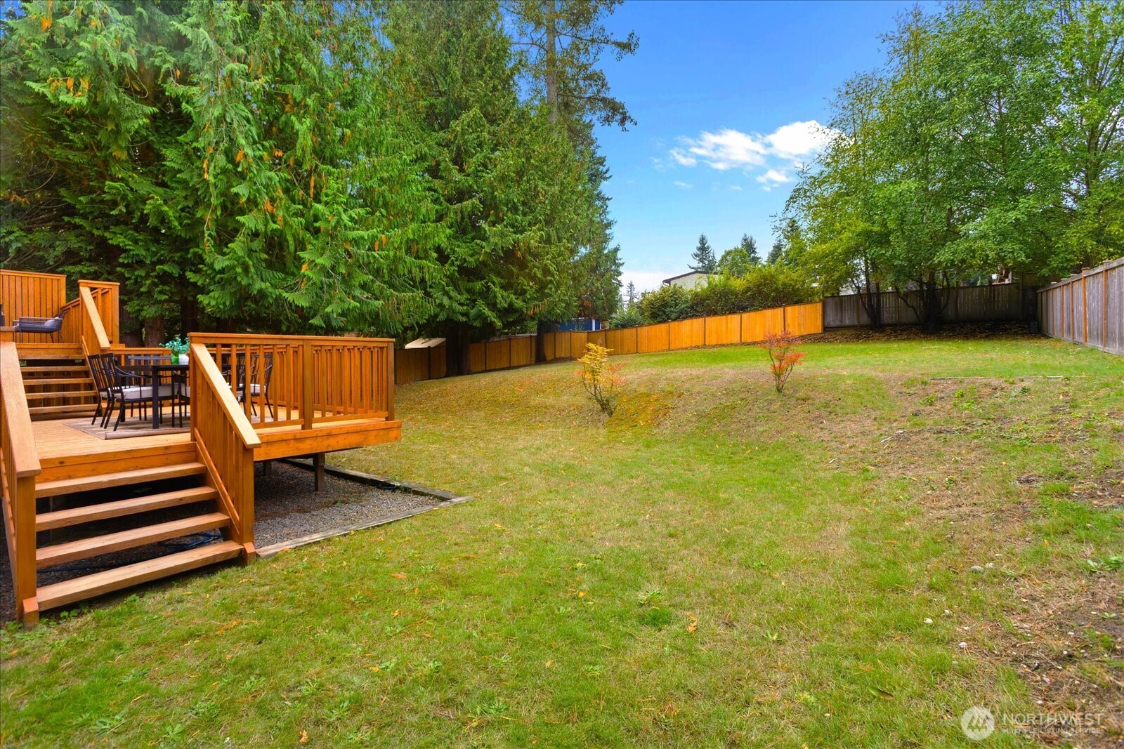 Fenced backyard with a broad lawn, mature trees, and a raised wooden deck with outdoor dining furniture.