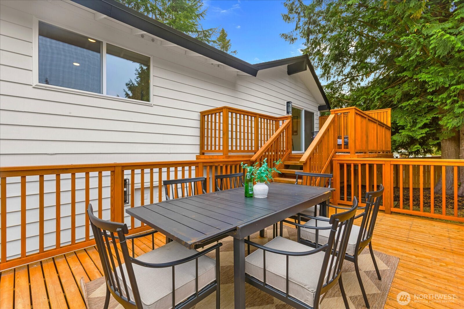 Furnished wooden deck with dining table and chairs beside a white-sided home.