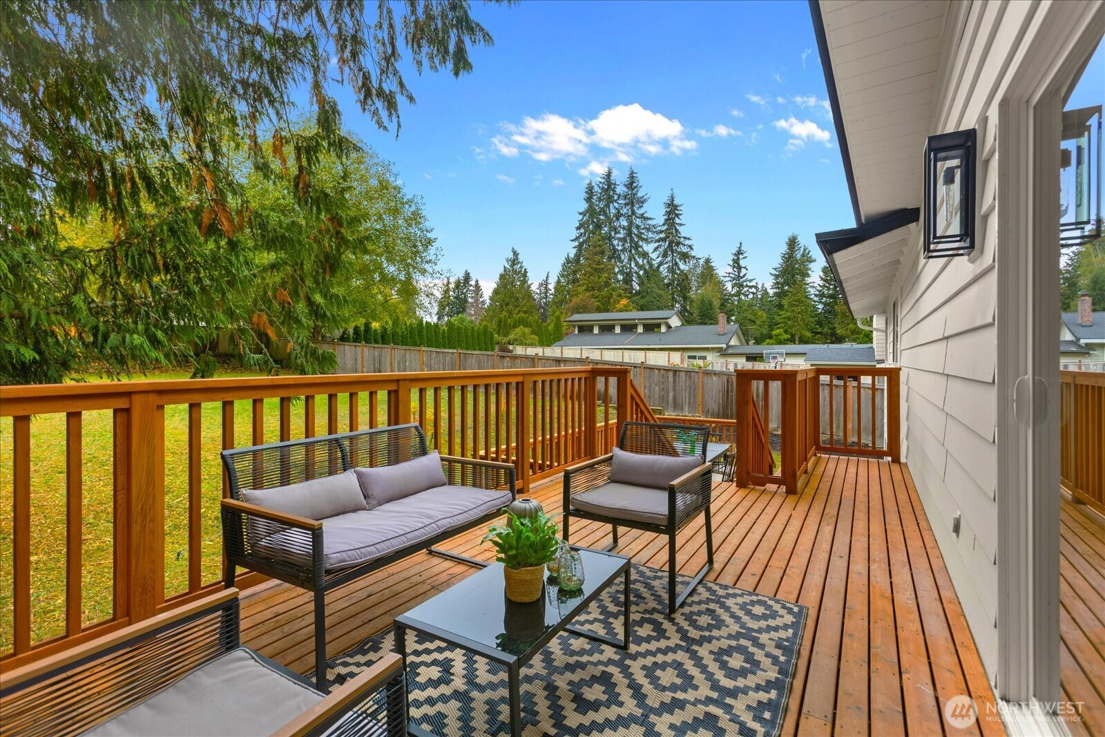 Furnished wooden deck with outdoor seating, a patterned rug, and a fenced, tree-lined yard.