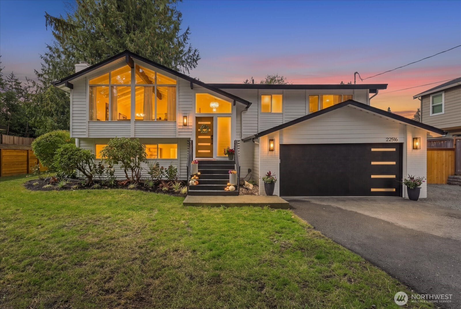 Modern gray split-level home at dusk with a peaked window, black garage door, and warmly lit windows.
