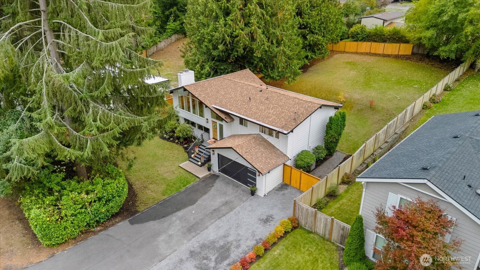 Aerial view of a white house with a fenced backyard, driveway, and mature evergreen trees.