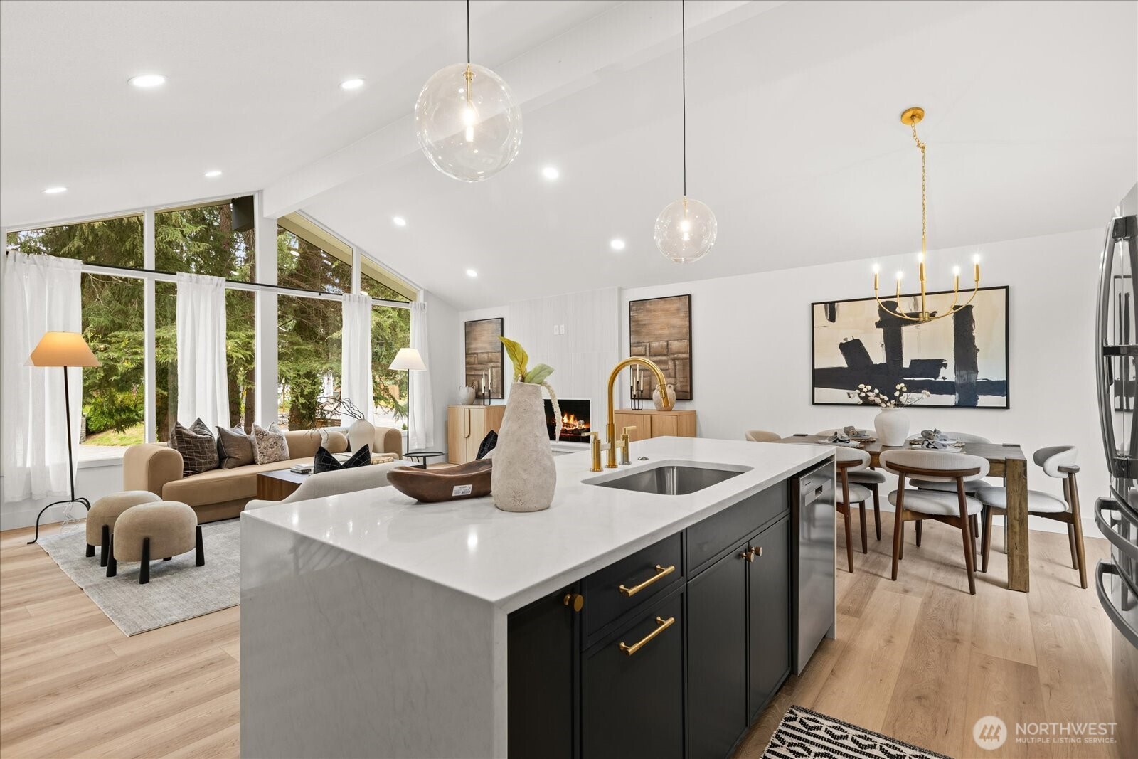 Open-plan kitchen with a white waterfall island, gold fixtures, vaulted ceiling, and tall windows.