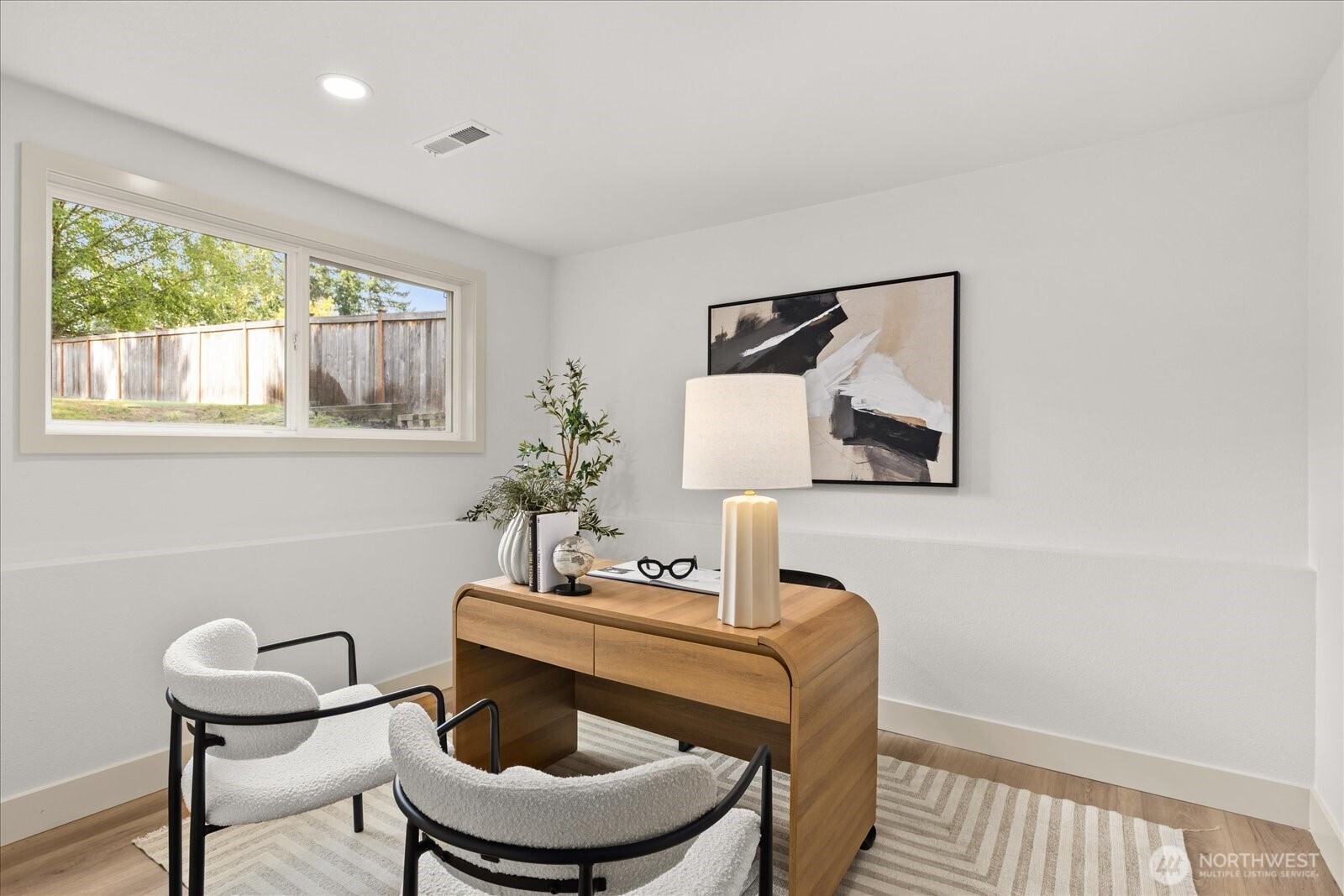 Furnished home office with a wooden desk, two white chairs, and a window overlooking a fenced yard.