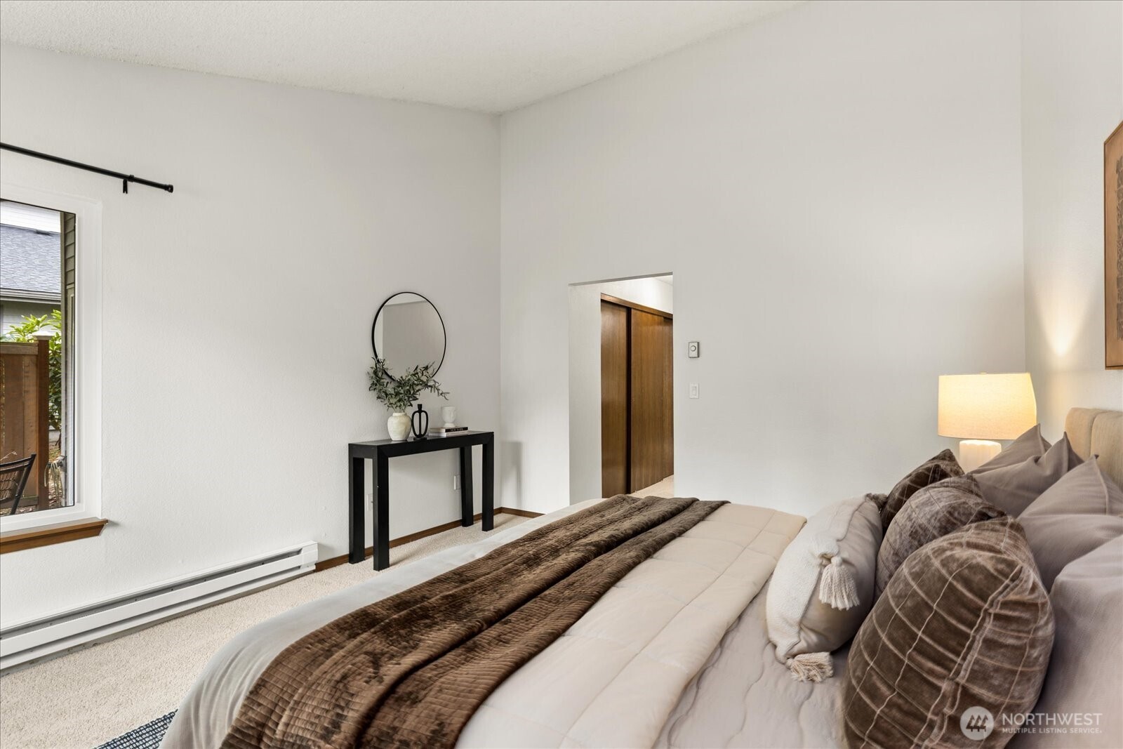 Staged bedroom with a white bed, brown quilt, taupe pillows, and black console table.