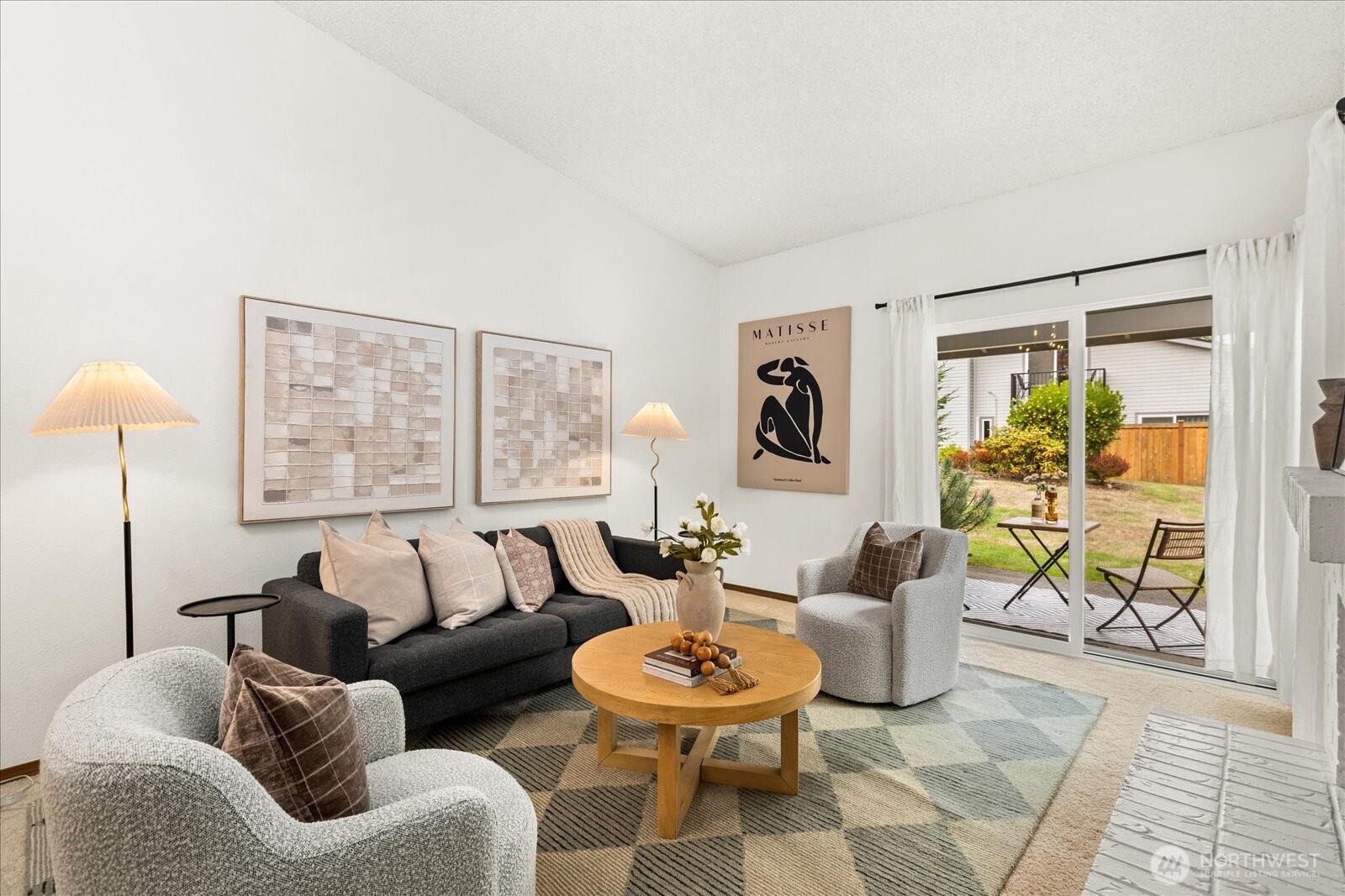 Staged living room with a charcoal sofa, pale armchairs, and sliding doors to a fenced patio.