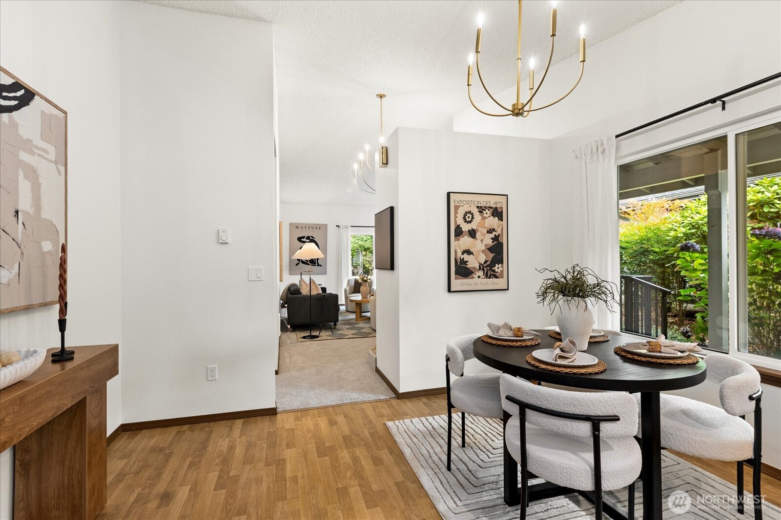 Dining room with a round black table, white upholstered chairs, and a sliding glass door overlooking greenery.